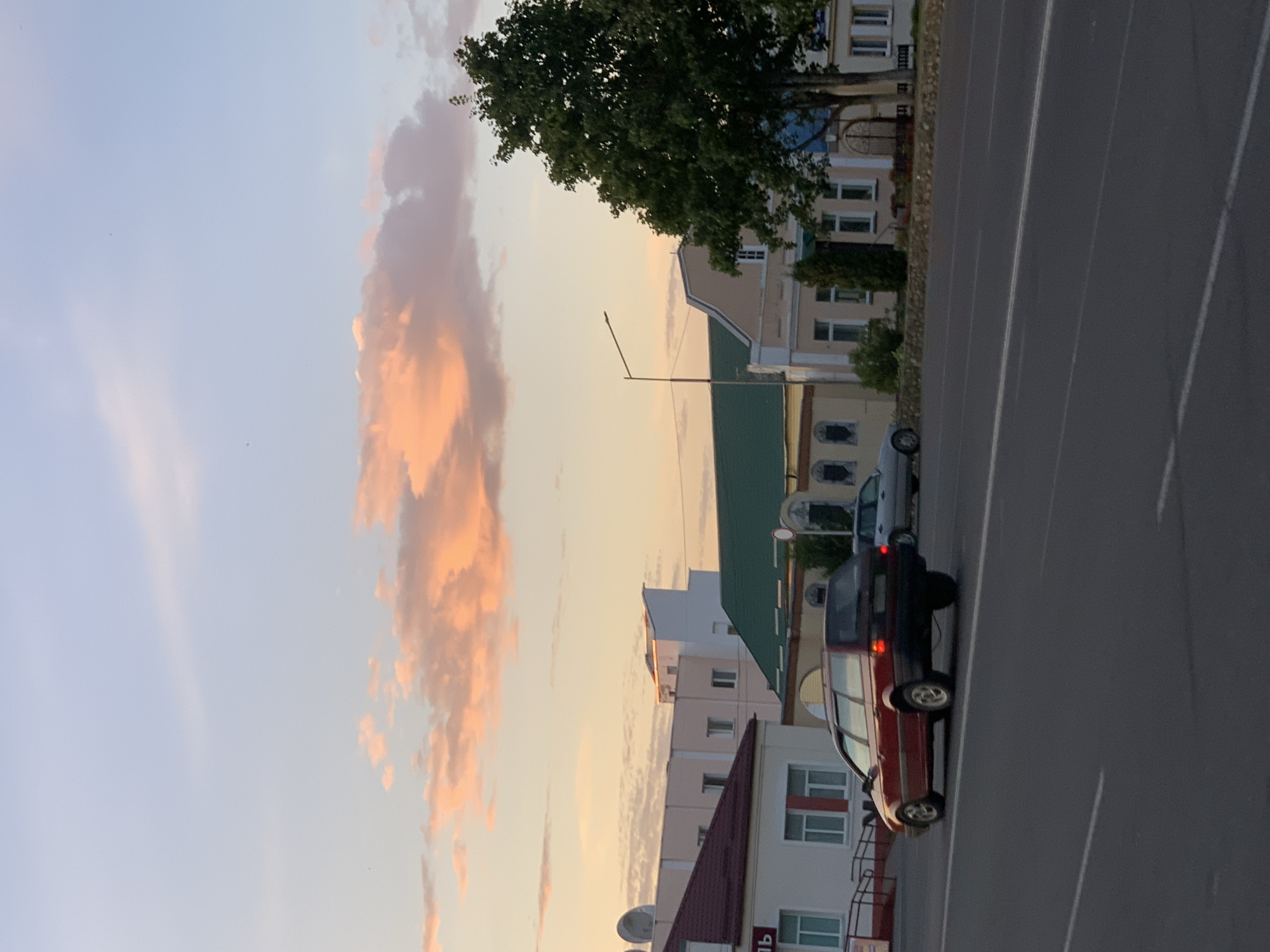 In Kapyĺ, Belarus, a dark red station wagon is captured driving on a multi-lane road during dusk. The sky displays a gradient of light blue to a soft orange, illuminated by the setting sun, with scattered clouds tinged with pink and orange. To the left of the road, several buildings are visible, including a structure with a red roof and white facade, and a larger building with a green roof. A large tree with dark green foliage stands on the right side of the road. The scene appears to be in a developed area, possibly a town or city.