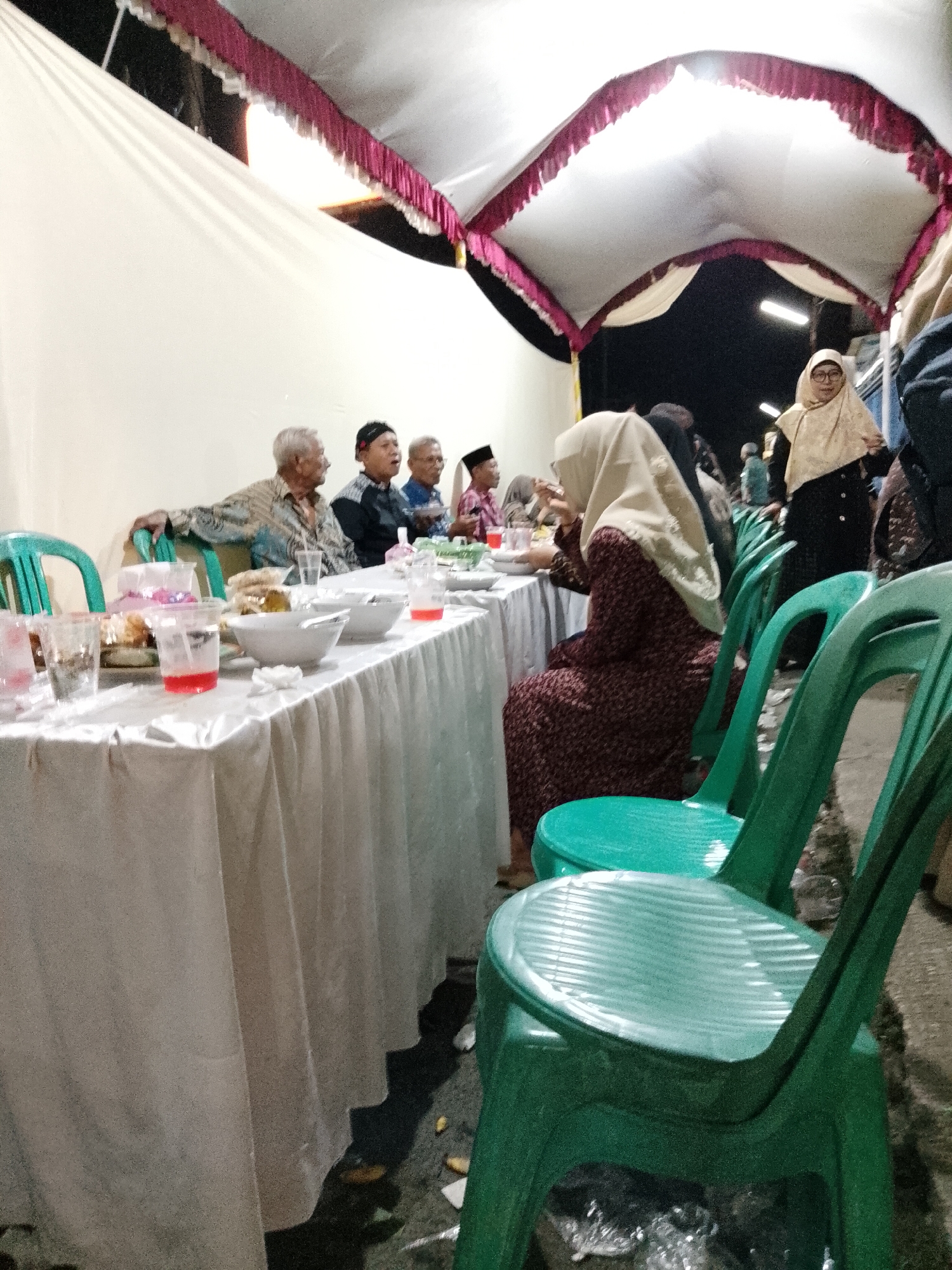 The image depicts an outdoor evening gathering under a draped tent in Trangkil, Indonesia. A long white table is set with white tablecloths, bowls, plates, and drinks in plastic cups. Several individuals are seated at the table, including three elderly men in traditional attire, and a woman in a hijab. In the foreground, two green plastic chairs are visible, and in the background, other people can be seen, including a woman in a light-colored hijab and black clothing, and a person in a dark top and pants. The lighting suggests the event is taking place at night, with artificial lights illuminating the area. The ground appears to be asphalt or concrete, with some debris scattered around. The overall scene suggests a communal meal or celebration.