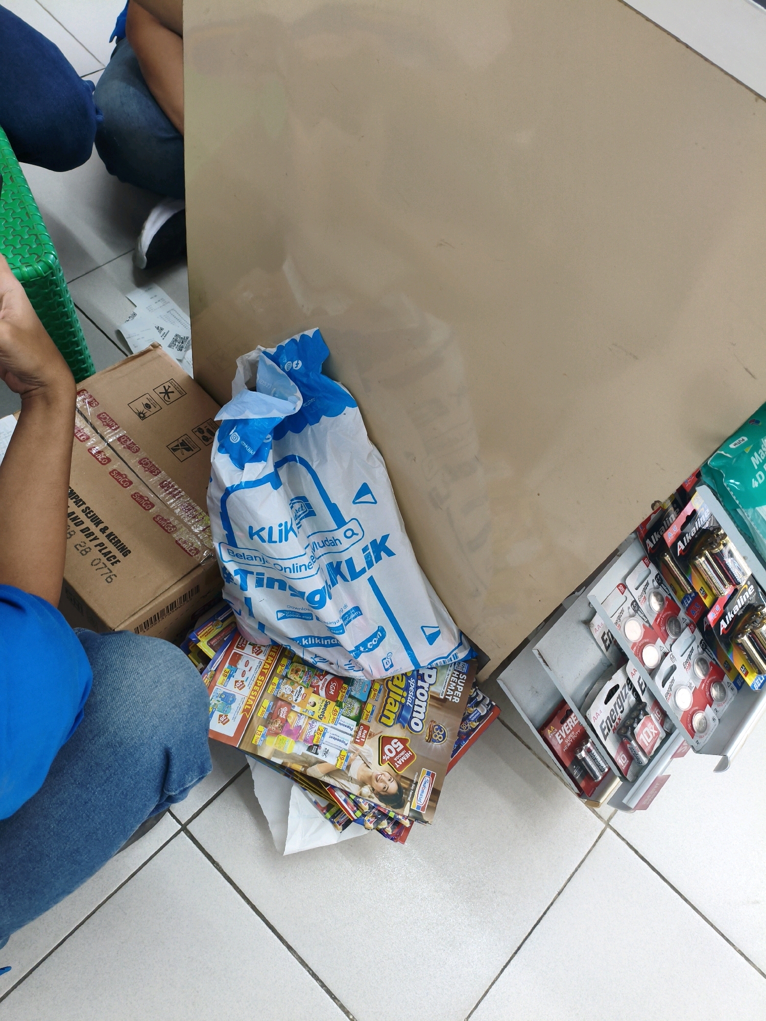 The image depicts a cluttered scene on a tiled floor, likely inside a store or storage area in Sumberejo Kulon, Indonesia. In the foreground, a person wearing blue jeans and a blue shirt is partially visible, sitting on the floor. To the right of the person, a white plastic bag with blue text and graphics, including the word "Klik," is prominently displayed. Beside the bag, several stacks of batteries in blister packaging are arranged in a metal display unit. The batteries appear to be of various sizes and brands, including Energizer. Behind the plastic bag and batteries, a cardboard box with text and graphics is visible. The box has tape with the "SunCo" logo. Scattered around the floor are promotional flyers or magazines, some with images of people and colorful text. The background consists of a large, light brown surface, possibly a wall or a piece of furniture. The overall impression is one of merchandise and packaging, suggesting a retail or distribution environment.