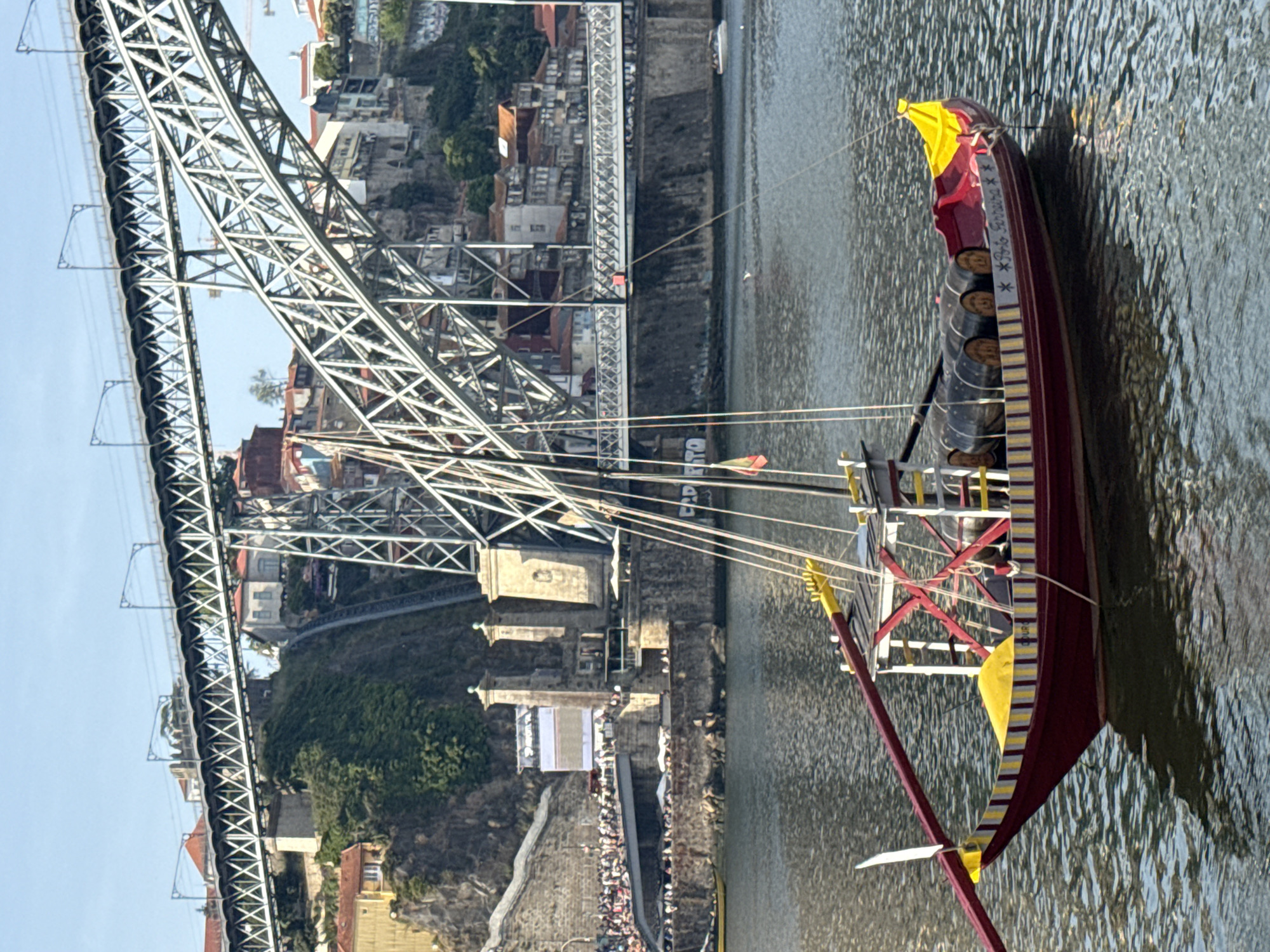 A rabelo boat, traditionally used for transporting port wine barrels, is docked on the Douro River in Vila Nova de Gaia, Portugal. The boat, painted red with yellow and white striped accents, carries several barrels. In the background, the Dom Luís I Bridge, an iconic metal arch bridge, spans the river. The city of Porto is visible on the opposite bank, with its colorful buildings lining the steep hills. The sky is clear and blue.
