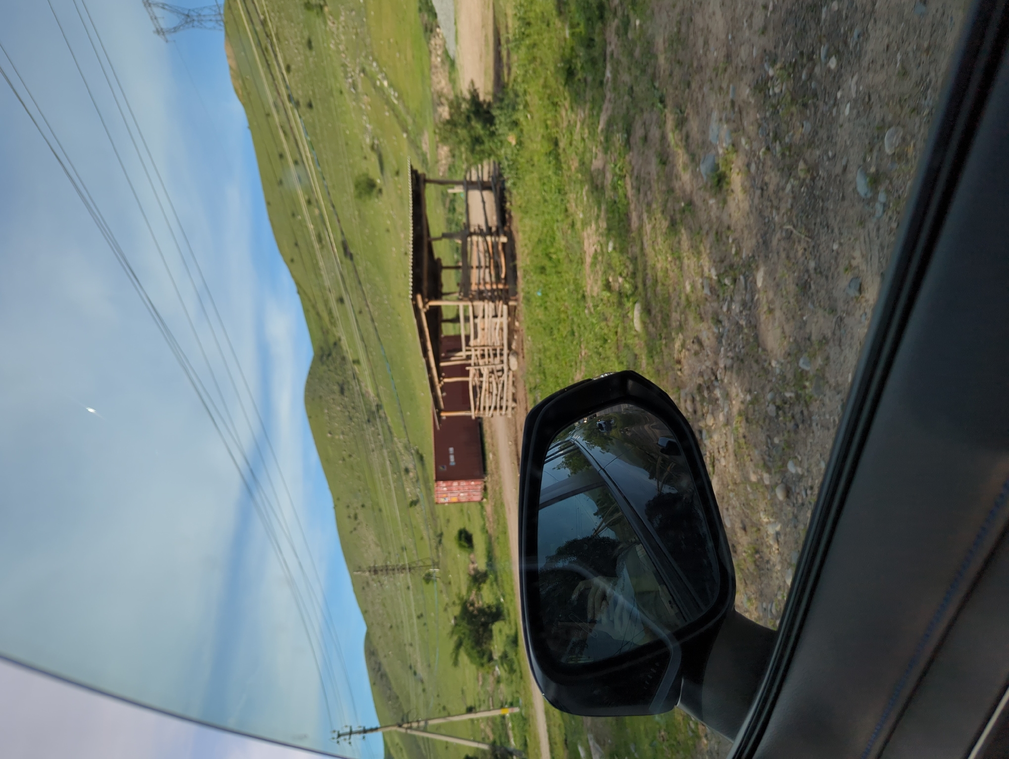 The image shows a view from inside a vehicle, looking out the passenger window. In the foreground, the car's side mirror and interior trim are visible. The mirror reflects a distorted view of the surrounding landscape.
In the background, a hillside covered in green vegetation ascends toward the sky. Power lines and pylons cut across the landscape. At the base of the hill, there appears to be a crude, open-sided wooden structure. Next to the structure is a shipping container. The ground near the structure is gravel and dirt.
The location is likely along a road in a rural area near Koi-Tash, Kyrgyzstan, given the mountainous terrain and basic infrastructure.