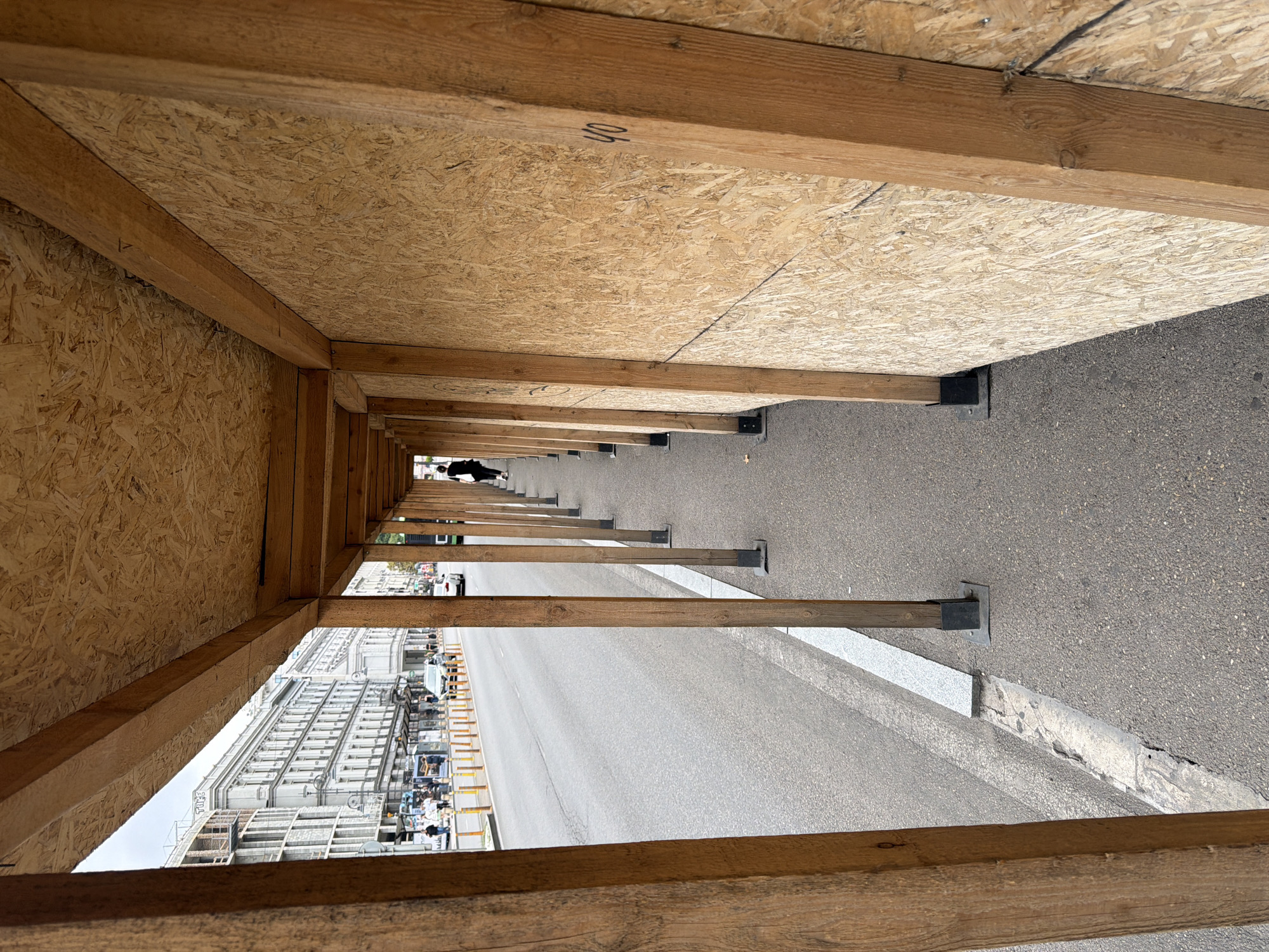 The image depicts a street scene in Tbilisi, Georgia, viewed from beneath a temporary wooden structure that appears to be a protective walkway or scaffolding. The structure is composed of vertical wooden posts and a roof made of oriented strand board (OSB). The walkway runs alongside a paved road with a white line marking the edge. On the other side of the road, there are buildings with multiple stories, some appearing to be old and ornate, suggesting an urban environment. Several vehicles and pedestrians are visible on the street. A lone figure is walking away from the camera within the covered walkway. The OSB panels forming the wall of the walkway have markings on them, including "40". The overall impression is that of an active construction or renovation site within a city.