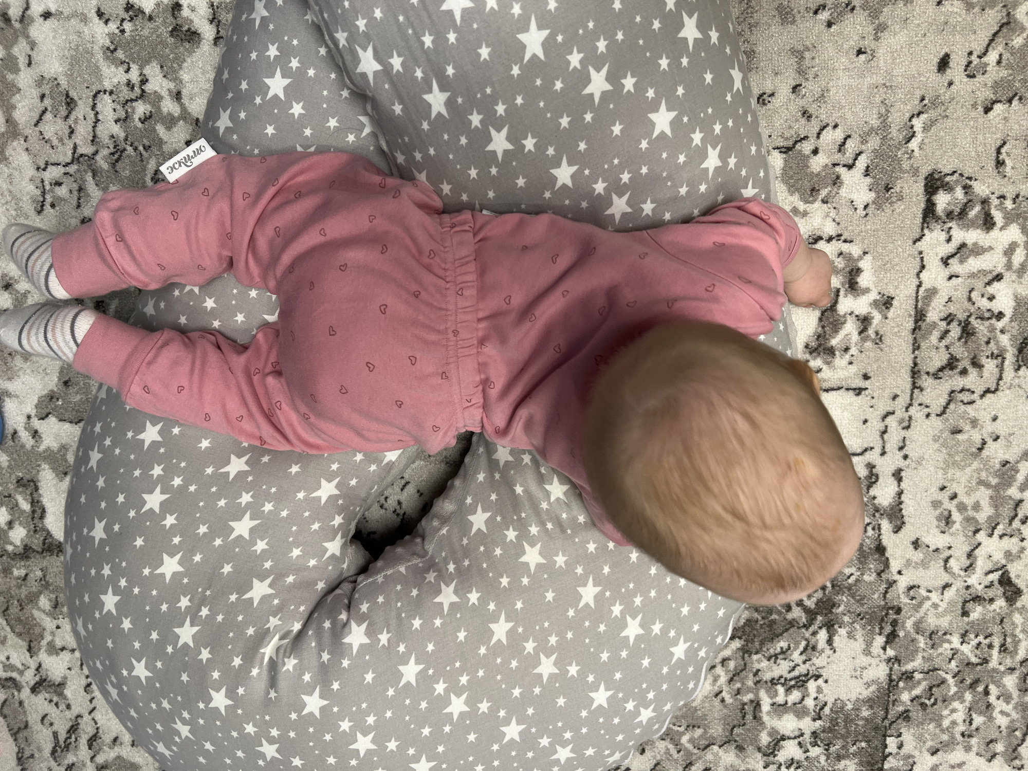 A baby, prone and wearing a pink onesie with small heart prints, rests on a gray and white star-patterned nursing pillow. The baby's blonde hair is visible at the top of the frame. The scene takes place on a gray and white patterned rug. A clothing tag with the word "эскимо" (Eskimo in Russian) is visible near the baby's left leg. The exact location within the city of Chaikovsky, Russia is not determinable from the image.