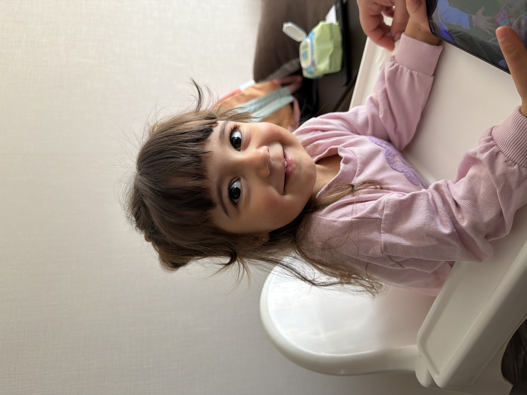 A young child sits in a white highchair, holding a black tablet in their hands. The child, with brown hair and eyes, is wearing a pink long-sleeved shirt. A pack of baby wipes is visible behind the child, resting on a brown couch. The background consists of a light-colored wall.