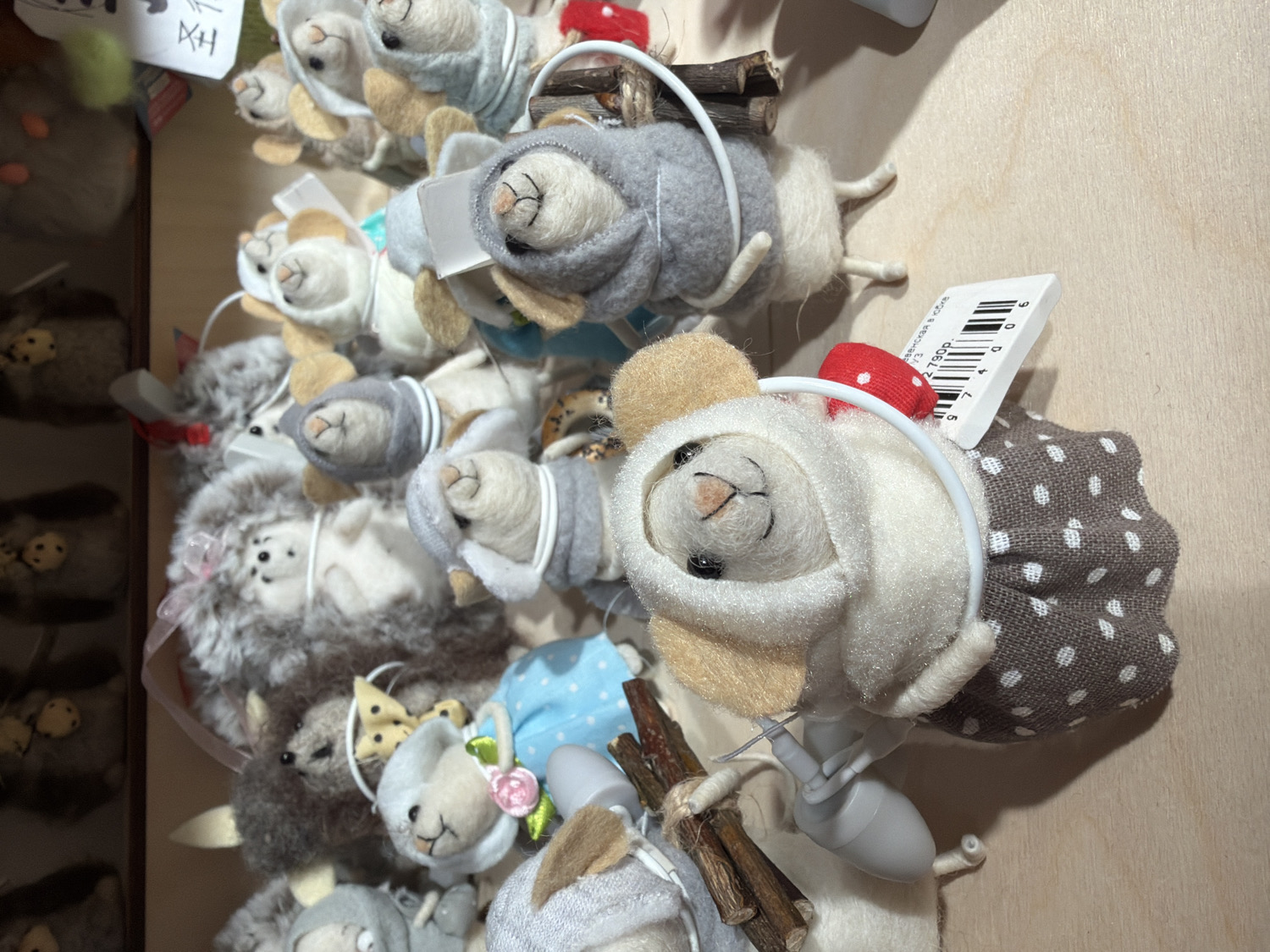 This media file presents a close-up, slightly overhead view of a vibrant display of numerous small, handcrafted felt or plush animal figures, likely for sale in a retail setting in Saint Petersburg, Russia. The subjects are primarily stylized mice and some hedgehogs, each meticulously designed with a whimsical and endearing aesthetic.
The figures are arranged densely on a light-colored wooden surface, which appears to be a display shelf or table. Each animal is uniquely adorned with miniature clothing and accessories. Examples include mice wearing grey sweaters, hooded cloaks, or patterned dresses (such as a brown dress with white polka dots or a blue polka-dot dress). Many figures feature tiny hats, like a bright red mushroom or delicate floral decorations, and some hold small bundles of sticks, adding to their charm. Their faces are simple, embroidered, and uniformly depict small, content smiles, contributing to their cute and innocent appeal. Some figures are suspended by thin white wires, suggesting they may be intended as ornaments or hanging decorations.
To the left, a darker, multi-tiered display area is partially visible, holding more similar items. The lighting is bright and even, characteristic of an indoor shop or market stall.
Visible text includes a white tag attached to one of the figures, displaying "2790P" and a barcode. Above the barcode, partial Cyrillic text reads "Упаковка 8 см" (Packaging 8 cm). Another tag in the upper left corner shows some illegible characters, possibly "XH". The overall scene evokes a sense of a charming, artisanal gift shop or craft fair.