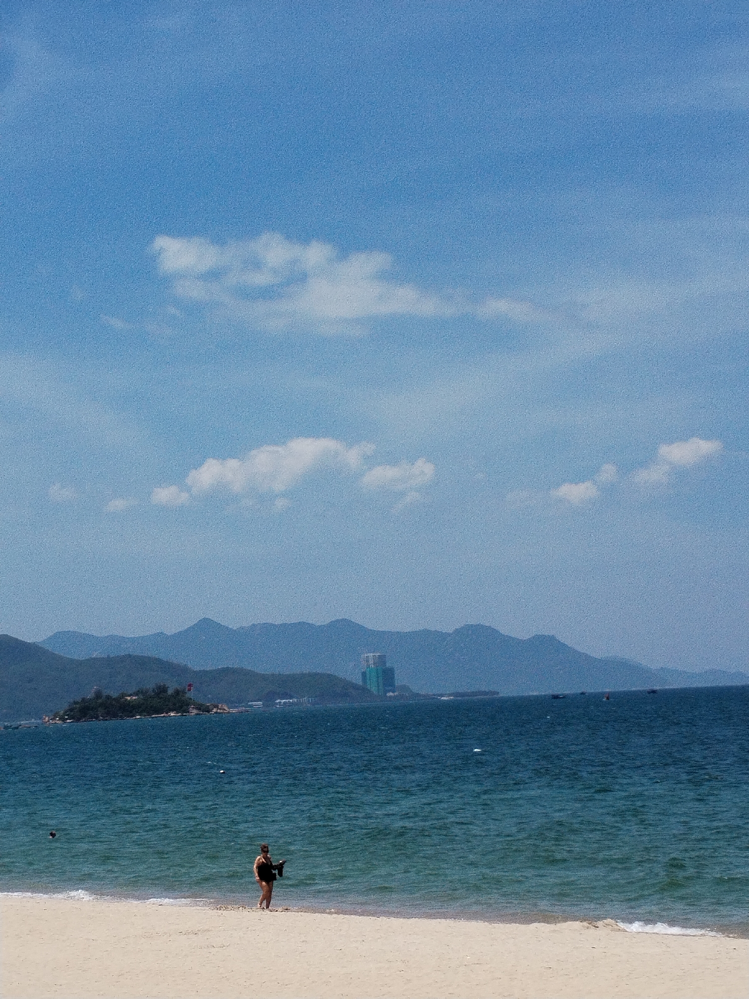 This is a bright, slightly grainy, full-frame, eye-level shot of a beach scene in Nha Trang Ward, Vietnam. The scene is dominated by a clear blue sky with a few scattered white clouds. In the foreground, a wide expanse of pale sand stretches to meet the turquoise ocean. A person wearing dark swimwear stands near the water's edge on the sand, holding a black object. The ocean exhibits gentle waves and a few buoys are visible. In the middle ground, a small island with trees and a structure topped with a red flag is situated off the coast. Beyond the island, a range of blue-hued mountains forms the background, with a tall, modern building visible on the far right behind the mountains. Several small boats are scattered across the water in the distance. The sun appears to be high in the sky, casting a bright light on the scene.