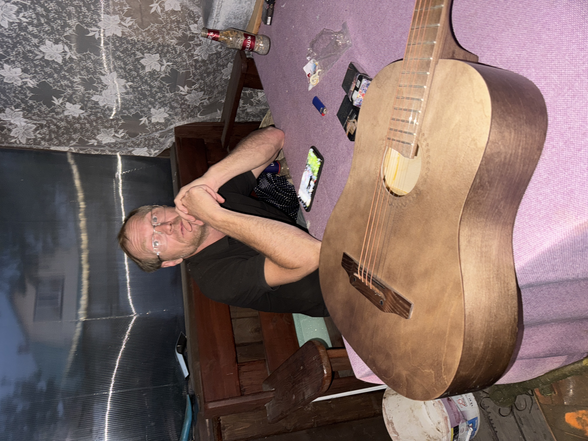 A man with glasses and thinning hair sits at a table covered with a pink tablecloth. He is wearing a black t-shirt. His hands are clasped in front of him, resting on his chin. In the foreground, a wooden acoustic guitar rests on the table. A bottle of "Edds" brand alcohol, two packs of cigarettes, a lighter, and a mobile phone are also on the table. Behind the man, a wooden bench and a corrugated plastic wall are visible. A floral curtain hangs to the right. The scene appears to be an outdoor, covered area, possibly a veranda or a gazebo, in Chaikovsky, Russia.