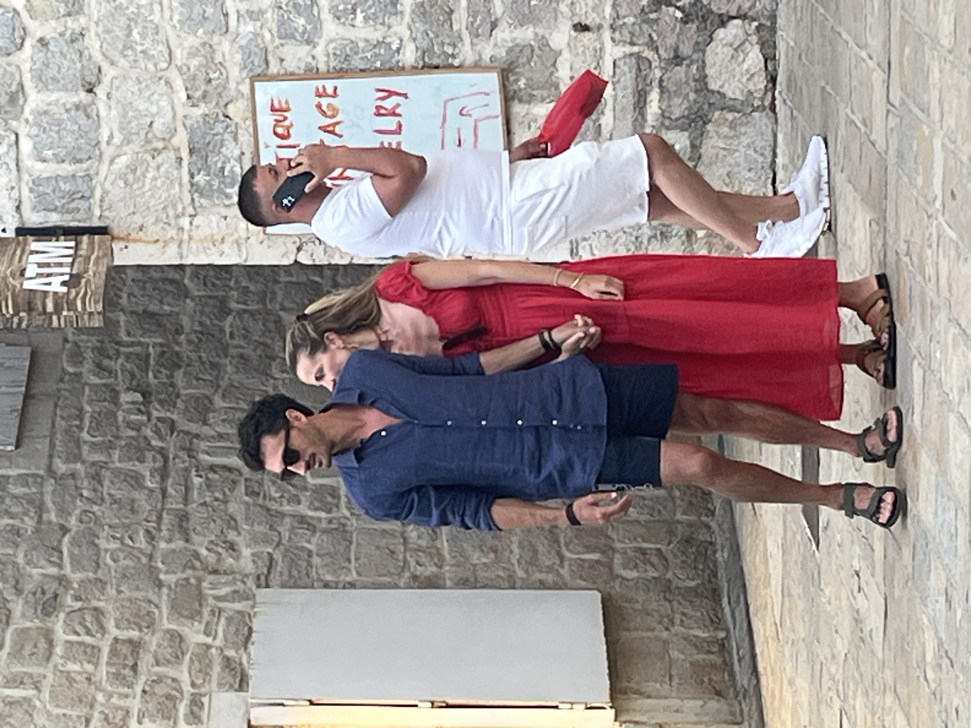 A man and a woman are standing close together on a cobblestone street in Budva, Montenegro, near a stone building. The man, on the left, wears a dark blue linen shirt and dark shorts, holding the woman's hand. He has dark hair, sunglasses, and a mustache. The woman, in a long red dress, stands beside him, also holding his hand. Further to the right, a man in a white t-shirt and white shorts is talking on a mobile phone, holding a red bag in his left hand. Behind him, a sign on the stone wall reads "ATM" and another sign has red writing, partially obscured, that appears to say "BOUTIQUE VINTAGE JEWELRY". The scene suggests a casual daytime activity on a European street.