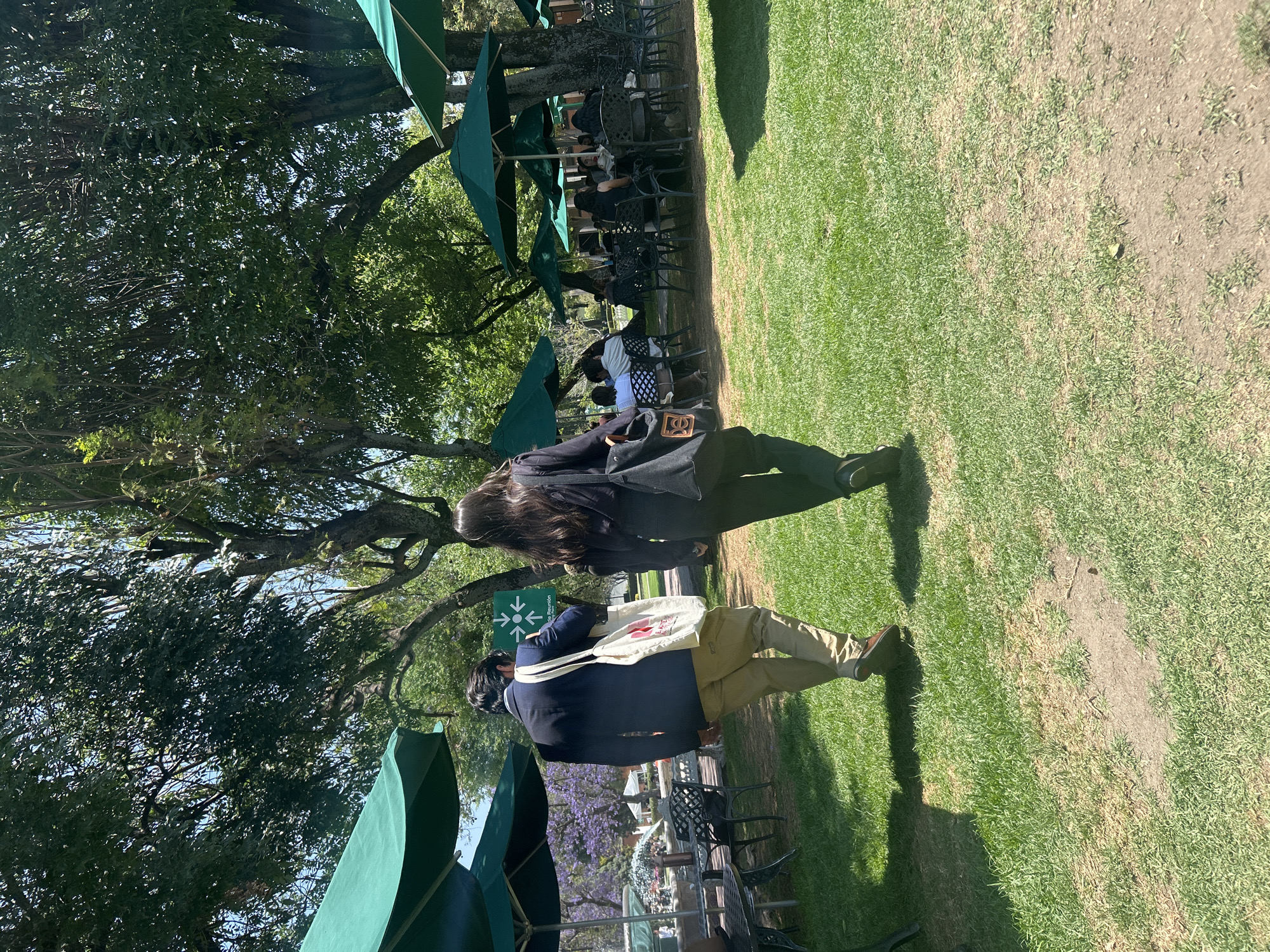 The image shows an outdoor scene in San Bernardino Tlaxcalancingo, Mexico, featuring a grassy area with trees and shaded seating areas. Two people are walking through the scene. One is wearing a dark blazer, tan pants, and carrying a white bag with red text and illustration. The second person is wearing a dark jacket and pants. There are numerous green umbrellas shading tables and chairs where other people are seated. Trees and other vegetation surround the area.