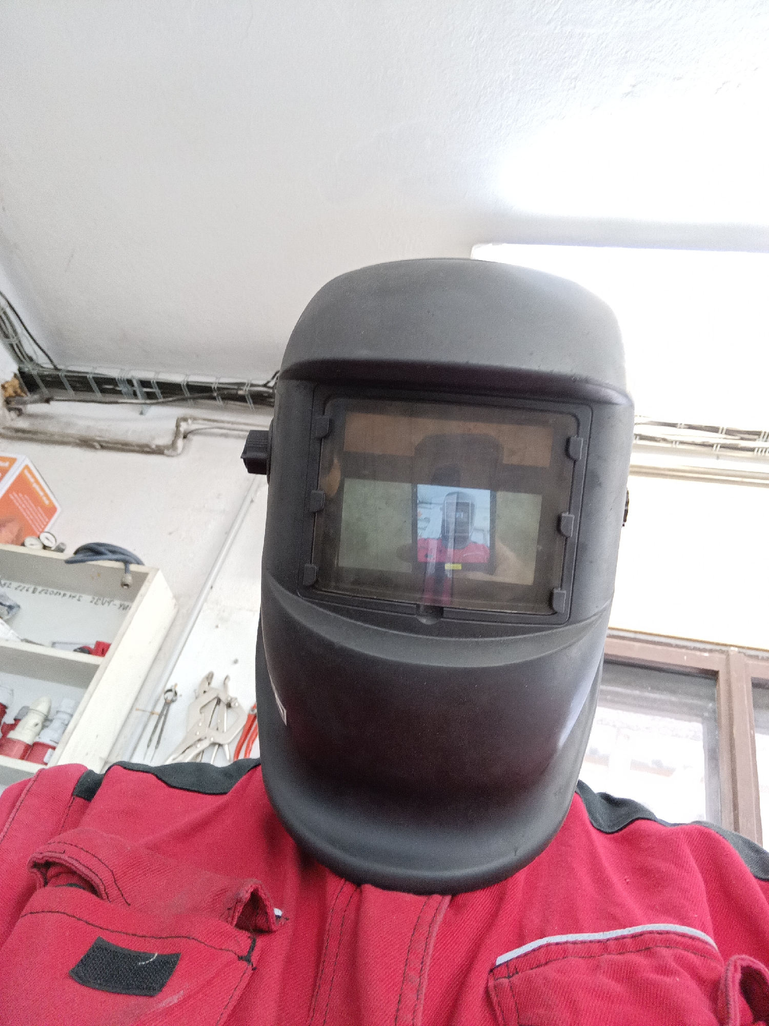 A close-up, eye-level, over-the-shoulder shot shows a person wearing a black welding mask and a red work jacket with black trim. The welding mask covers the majority of the face, with a tinted viewing window that reflects the person's face and upper body, wearing a red shirt. The jacket has visible stitching and a black patch on the left chest area. Behind the person, a white ceiling with visible electrical conduits and lighting fixtures can be seen. To the left, a white shelf unit contains various tools and supplies, including clamps and electrical components. A window with a wooden frame is visible to the right. The scene appears to be an indoor workshop or garage.