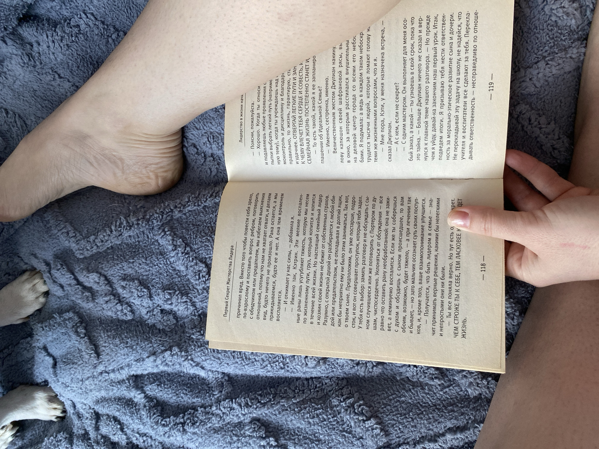 Here is a detailed description of the image:
**Scene:** The image shows a person lying on a gray textured blanket while reading a book. The book is opened to a two-page spread, revealing columns of text in Russian. The left-hand page is numbered "118," and the right-hand page is numbered "119." The book seems to be a self-help or leadership-focused book, given the titles “Первый Секрет Мастерства Лидера” and “Лидерство в жизни начи…” at the top of the pages.
**People and Subjects:**
- A person is lying on the blanket, holding the book open.
- The person's leg and foot are visible.
- The paws of a dog are partially visible in the lower left corner of the frame.
**Objects:**
- Book with Russian text
- Gray textured blanket
- Human leg and foot
- Dog's paws
**Actions and Interactions:**
- The person is reading the book.
- The dog is lying nearby, potentially interacting with the person.
**Environment:**
- The environment seems to be indoors, likely in a casual setting such as a living room or bedroom given the blanket.
**Location:** It is not possible to determine the city based on the visual elements in the picture. The image includes the leg and foot of the person reading the book.