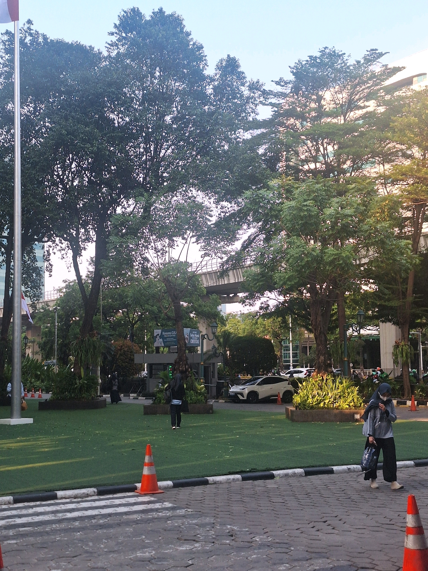 The image depicts a daytime scene in South Jakarta, Indonesia, featuring a wide paved area with a pedestrian crossing in the foreground. Two orange traffic cones with white stripes are visible on the paved area. Beyond the crossing, a large expanse of artificial turf lawn is present, dotted with traffic cones and planters. Several mature trees with dense foliage are scattered across the lawn and surrounding areas, with one prominent tree in the center casting shadows. In the midground, a white car is parked, and several people, including a woman wearing a hijab, dark pants, and a light-colored jacket, walk across the paved area. In the background, a modern building with large windows and an elevated railway track with a train overhead are visible. The sky is clear and bright, suggesting a sunny day.