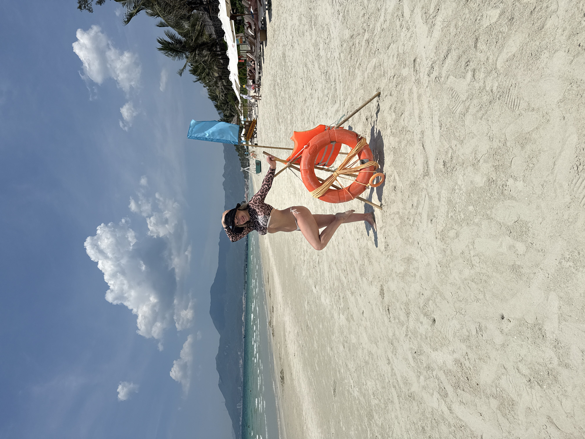The image depicts a woman posing on a sandy beach in Dong Ninh Hoa Ward, Vietnam. She is wearing a leopard-print swimsuit and a hat, and standing next to a makeshift structure made of sticks that holds a blue flag and two orange life preservers. In the background, there are palm trees, other beachgoers, mountains, and a sky with clouds. The tide appears to be receding, revealing a wide expanse of sand.