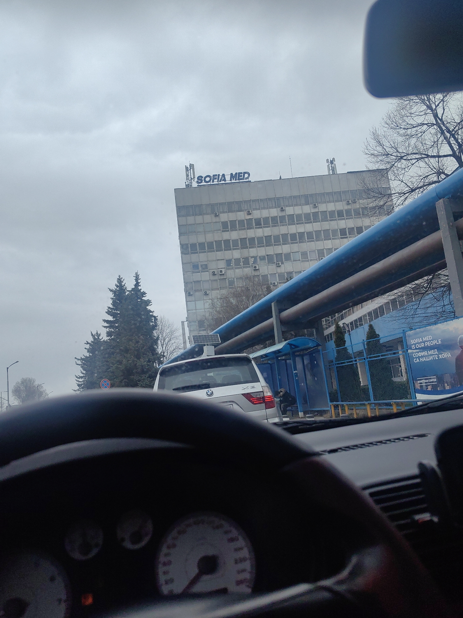 A photograph taken from inside a car looking out shows the Sofia Med hospital building in Sofia, Bulgaria. A silver BMW SUV is in front of the car, and a bus stop is on the right. Large blue pipes run along the front of the hospital. A billboard near the bus stop reads "SOFIA MED IS OUR PEOPLE" in both English and Bulgarian. The sky is overcast, and bare trees are visible in the background. The car's dashboard and steering wheel are visible in the foreground, out of focus.