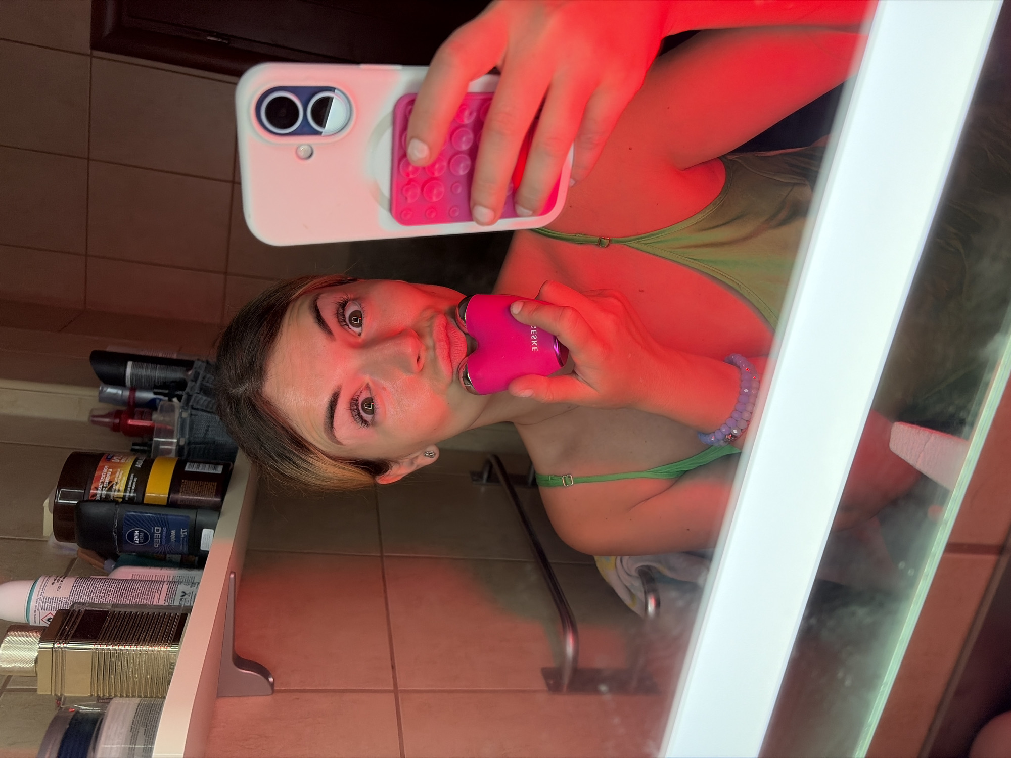 A woman is taking a selfie in a bathroom. She is holding a pink heart-shaped device to her mouth with her left hand and a smartphone in her right hand, which is angled to capture her reflection. The bathroom is tiled with brown tiles, and a shelf above the mirror holds various toiletries, including bottles and jars. A red light is illuminating the scene, casting a warm glow on the woman and her surroundings. The mirror reflects the woman and the bathroom behind her.