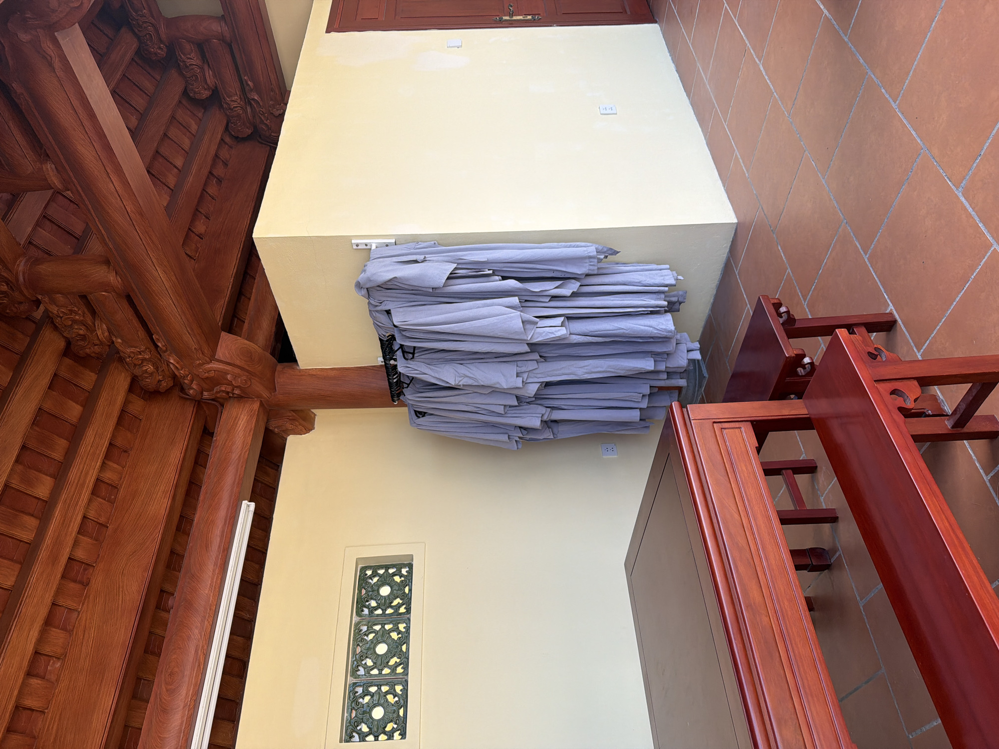 The image depicts an interior view, likely a temple or traditional building, situated in Nam Nha Trang Ward, Vietnam. Predominantly, a large pile of folded gray cloths or robes rests on a raised platform or ledge attached to a pale yellow wall. Above the cloths is a darker wood cabinet built into the wall. The wall is interrupted by a decorative, vertical window composed of green and white geometric patterns. Elaborate, dark-red-stained wooden beams form the ceiling and structural supports on the left side of the image, showcasing traditional Vietnamese architecture. Brown terracotta tiles make up the floor on the right side of the image. A piece of furniture, possibly a table or cabinet, made of the same dark-red-stained wood as the beams, is visible in the foreground on the right. The overall impression is one of a space undergoing preparation, perhaps for a religious ceremony or gathering. The image is well-lit, providing a clear view of the objects and architectural details.