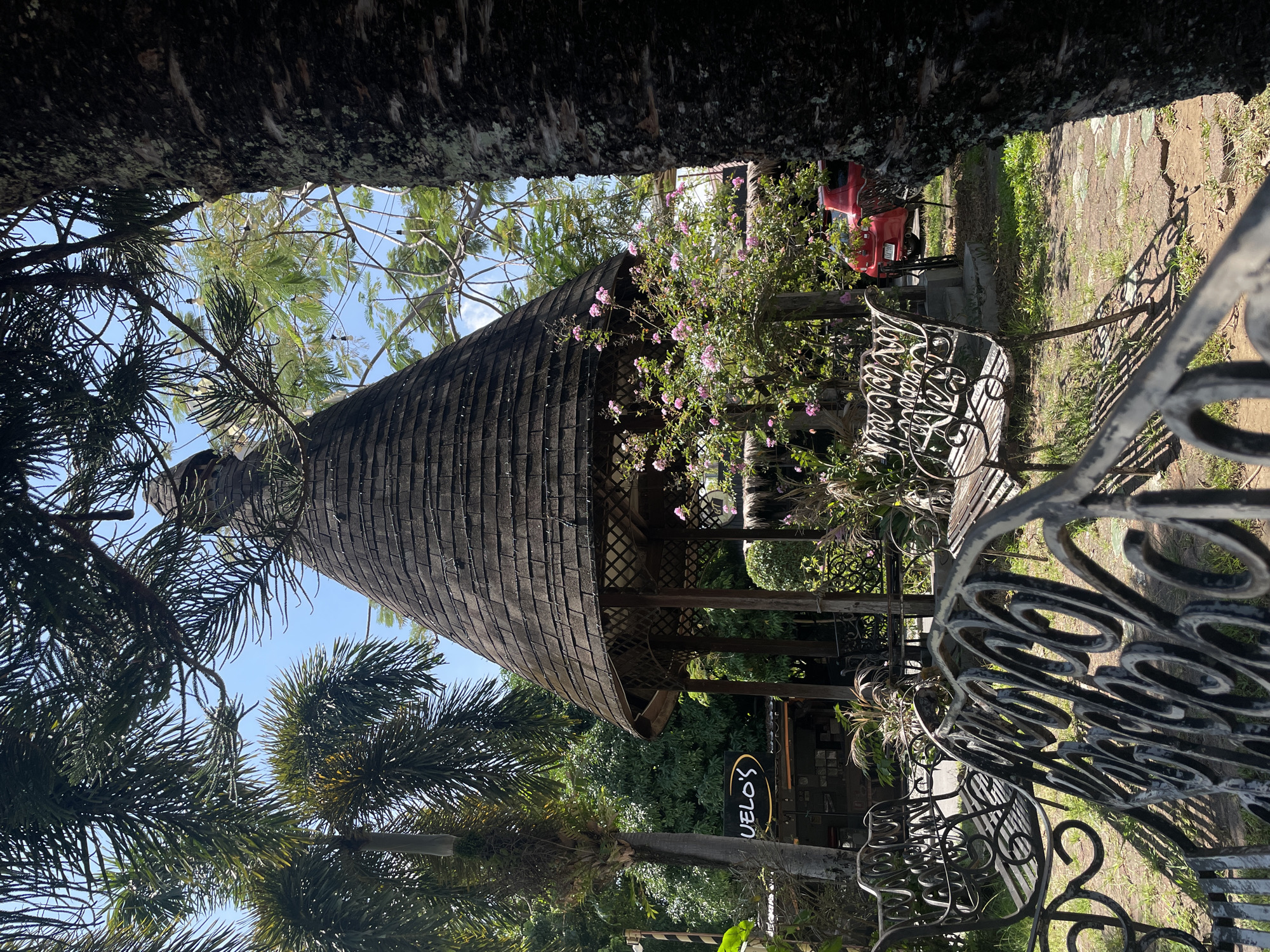 The image shows a gazebo with a conical, shingled roof, set in an outdoor area with lush green foliage and palm trees. Two ornate black metal benches are in the foreground. In the background, a red car is partially visible. A sign for "UELO'S" is also visible on a building. The scene suggests a relaxed, possibly park-like or restaurant setting. Based on the presence of palm trees and the overall tropical ambiance, this location could be in Davao City, Philippines, known for its abundant green spaces and warm climate.