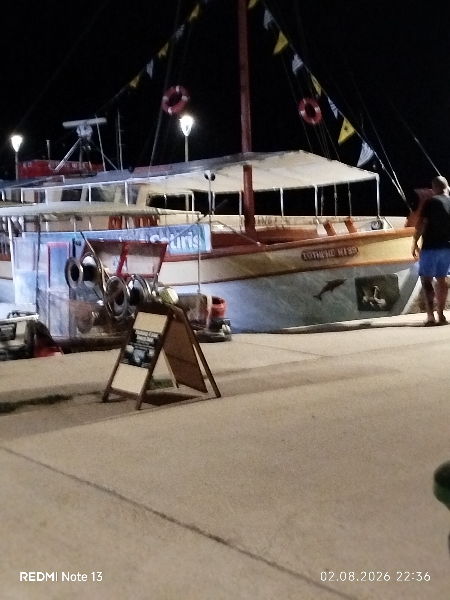 A night-time scene depicts a boat docked in a harbor. The boat, painted with a light blue hull and a tan superstructure, features a banner that appears to say "Saturis". Two life preservers, one red and one yellow, are visible on the boat's upper deck, along with various flags strung between poles. A person in dark shorts and a black shirt stands on the dock to the right of the boat. In the foreground, a wooden A-frame sign displays information. The ground is paved with concrete, and the overall lighting suggests it is after sunset. The text overlay at the bottom indicates the image was taken on August 2, 2026, at 22:36, and was taken with a REDMI Note 13. While the specific location in Sithonia Municipal Unit, Greece, cannot be definitively determined from the image alone, the setting is consistent with a coastal town or port in that region, possibly during a summer evening.