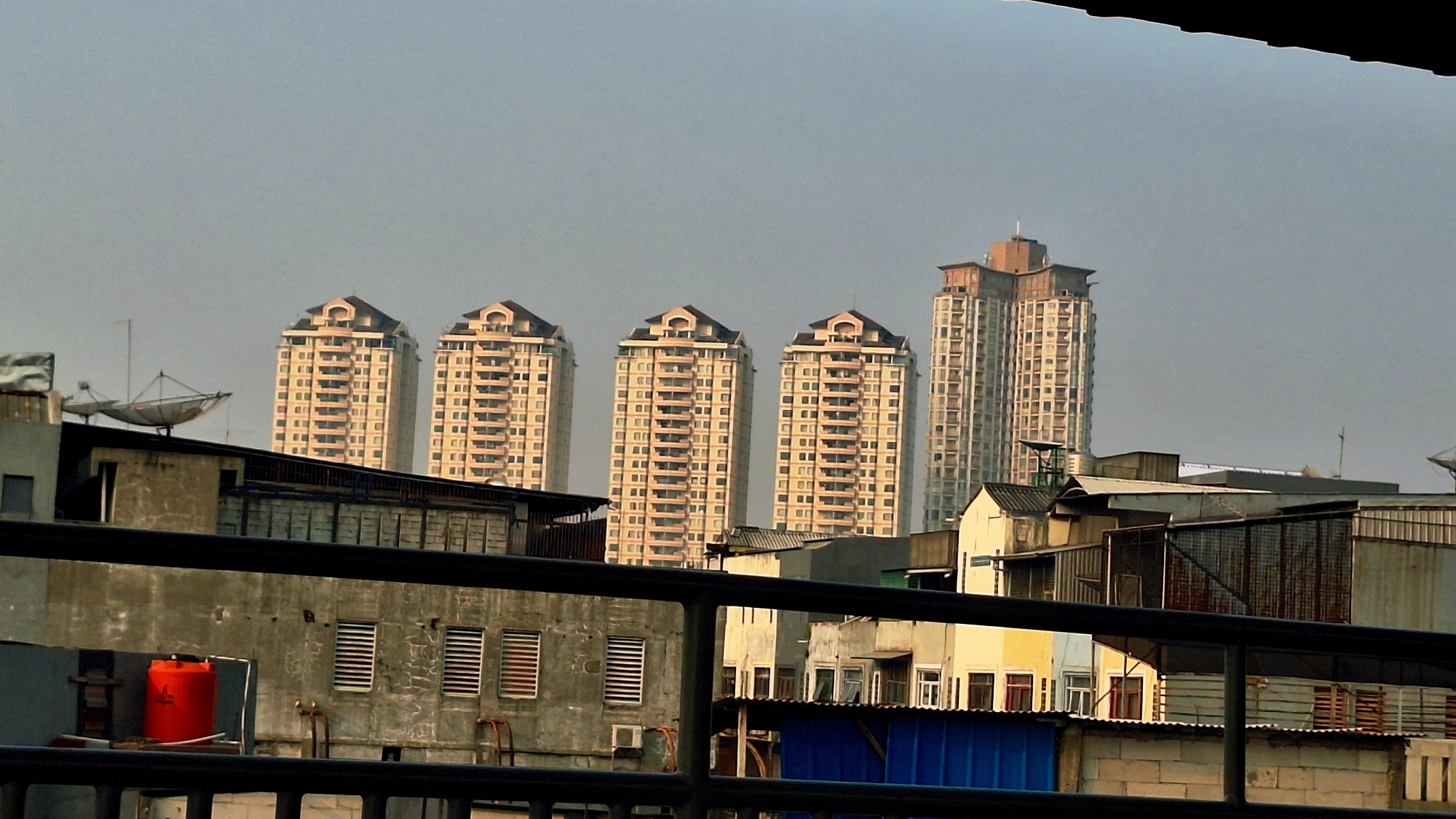 The image depicts a cityscape in North Jakarta, Indonesia, characterized by a dense collection of buildings under a hazy, overcast sky. In the background, a row of five identical, light-colored high-rise apartment buildings stands prominently, each featuring a distinctive stepped roofline. To the right of this row, a taller, more modern skyscraper with a visible construction phase is situated. In the foreground, a variety of lower-rise structures are visible, including what appears to be a concrete building with striped windows and a bright orange water tank on its roof. Several satellite dishes are mounted on some of the foreground buildings. The overall scene suggests an urban environment with a mix of residential and commercial architecture, possibly captured during sunrise or sunset given the muted light. There are no people or discernible actions captured in the image.