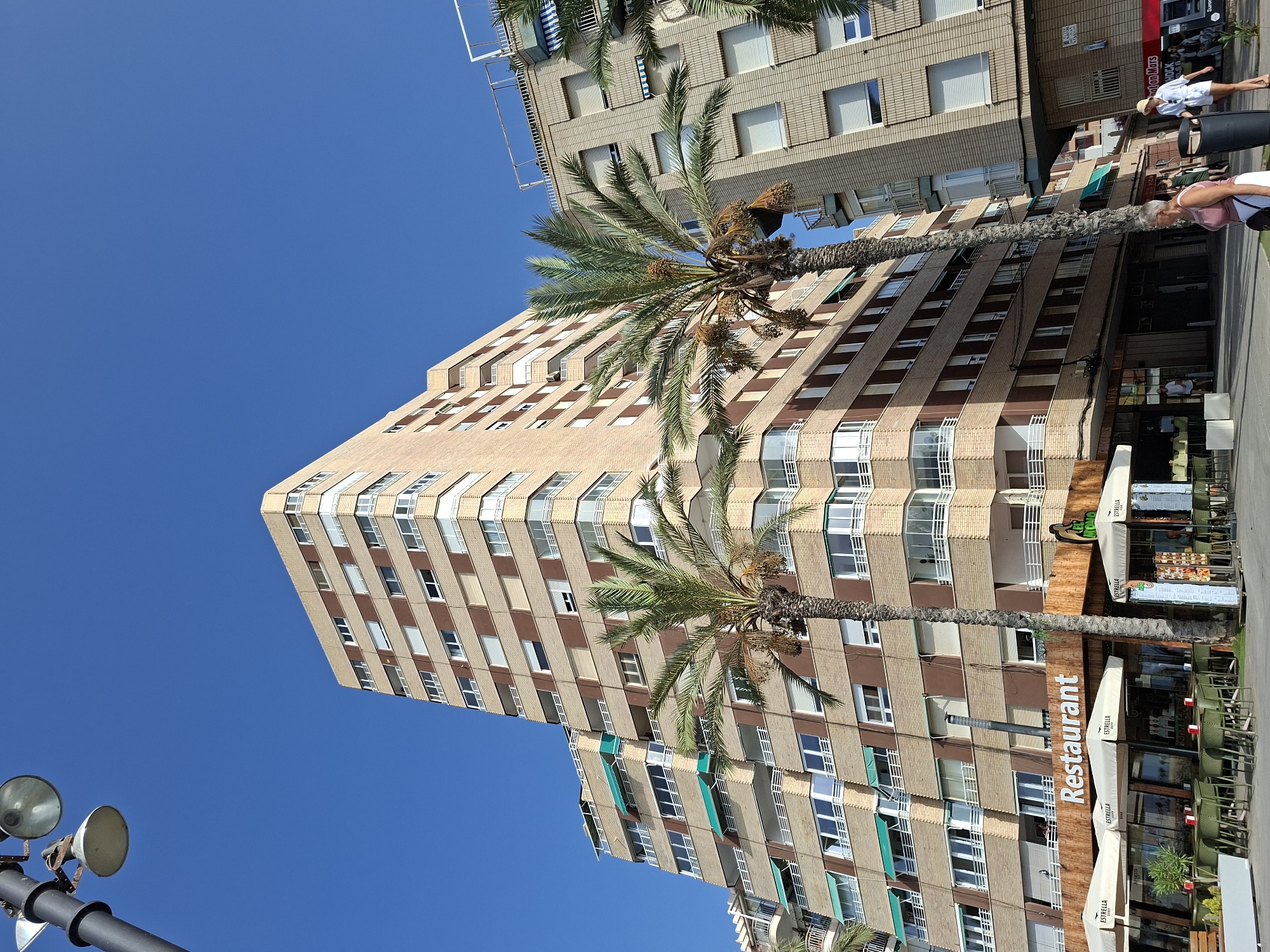 The image depicts a view of a street in Torrevieja, Spain, with a focus on buildings and pedestrian activity. Dominating the scene is a tall, multi-story building with a beige facade and brown accent panels, featuring numerous windows and balconies. To the right, another similar building is visible, also with a beige brick facade. Two palm trees are positioned in front of the buildings, one taller and fuller than the other.
At the ground level of the main building, there is a restaurant with outdoor seating. Several tables and chairs are set up under white umbrellas with the logo "ESTRELLA". The restaurant has a wooden facade with a prominent "Restaurant" sign in white lettering. Next to the restaurant, a menu board is displayed.
On the sidewalk, several people are walking. A woman in a pink top and white skirt is walking towards the left, with a dark handbag. Another woman in a white outfit and a straw hat is walking towards the right, barefoot. A man in a white shirt and shorts is walking further down the street to the left. The environment is characterized by a clear blue sky and sunshine, indicating a warm day. The street appears to be paved, and there is a trash bin on the sidewalk. There is also a street lamp visible in the upper left corner of the image.