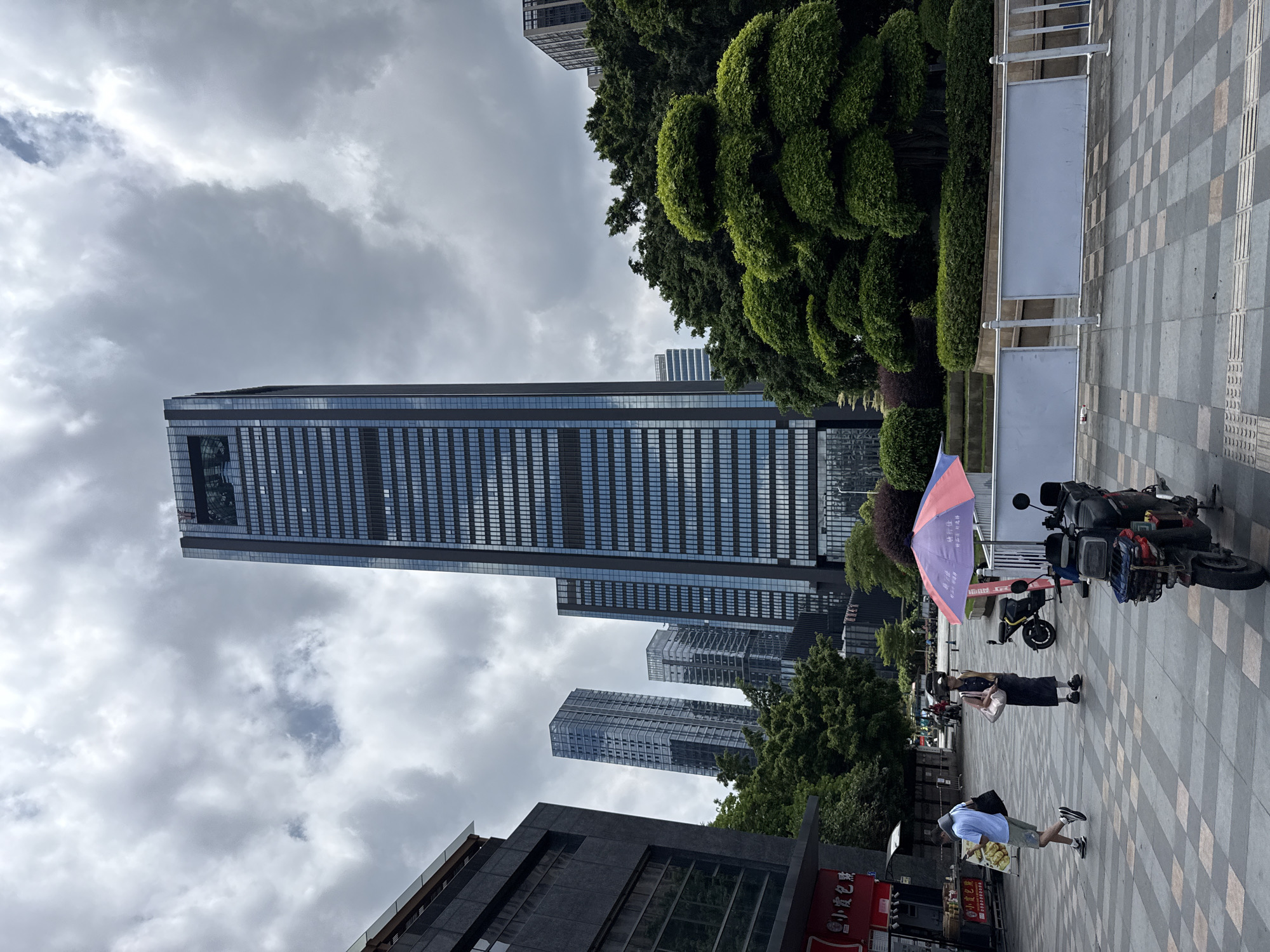 The image depicts a street scene in Haizhu District, China, characterized by a cloudy sky and a juxtaposition of modern architecture and greenery. In the foreground, a paved pedestrian walkway with a checkered pattern runs across the scene. Two individuals are walking on the walkway. One person, wearing a light blue shirt, shorts, and carrying a black bag, walks away from the camera, looking at their phone. Another person, wearing a dark dress and a hat, walks towards the camera carrying a pink bag. A pink and blue umbrella is set up on a stand. Two electric scooters are parked in the middle ground. A tall, modern glass-fronted skyscraper dominates the background, reflecting the cloudy sky. Behind the skyscraper, other buildings and lush green trees are visible, including artfully manicured bushes. The scene conveys a sense of urban life under a subdued sky.