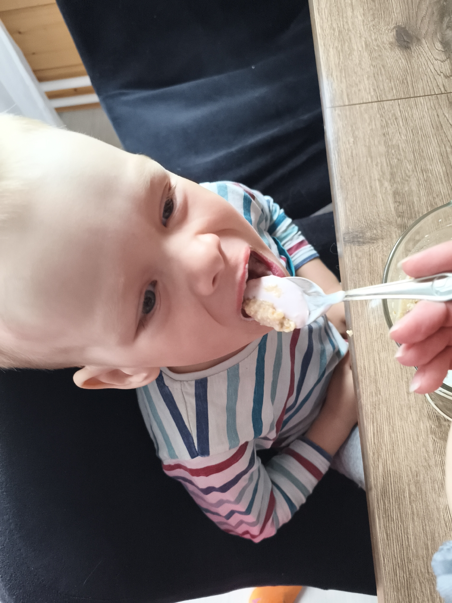 A young child with blond hair is being fed from a spoon by an unseen person. The child is wearing a striped shirt and is leaning forward, with their mouth open to receive the food. The food on the spoon appears to be a soft mixture of light brown and pink. The child's eyes are open, and they seem engaged in the activity. The setting appears to be indoors, with a wooden table surface visible. The child is seated, and a dark blue fabric, possibly clothing or furniture, is in the background. The image is taken from a slightly elevated angle, looking down at the child. The overall impression is one of a domestic mealtime. There is no visible text in the image.