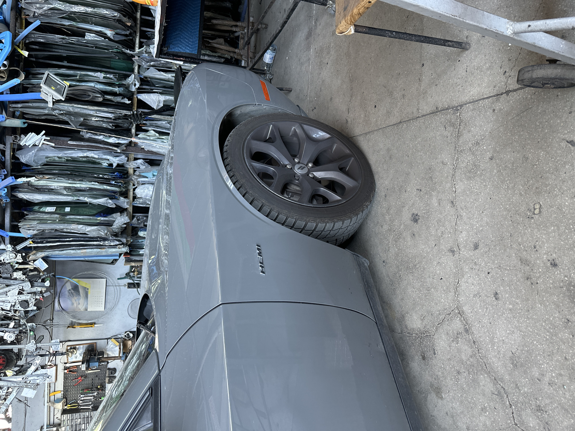 The image shows the side of a gray car, likely a Dodge Challenger, parked indoors in what appears to be an auto repair shop or a storage facility. The car's front passenger-side fender and wheel are visible. The vehicle has a "HEMI" badge on the fender. The background is filled with shelves stacked with various car parts, including windshields wrapped in plastic. Tools and equipment, such as a vise and a calendar, are mounted on a pegboard on the wall. The floor is concrete with cracks and some debris. A metal cart and a wooden stool are also present in the scene.