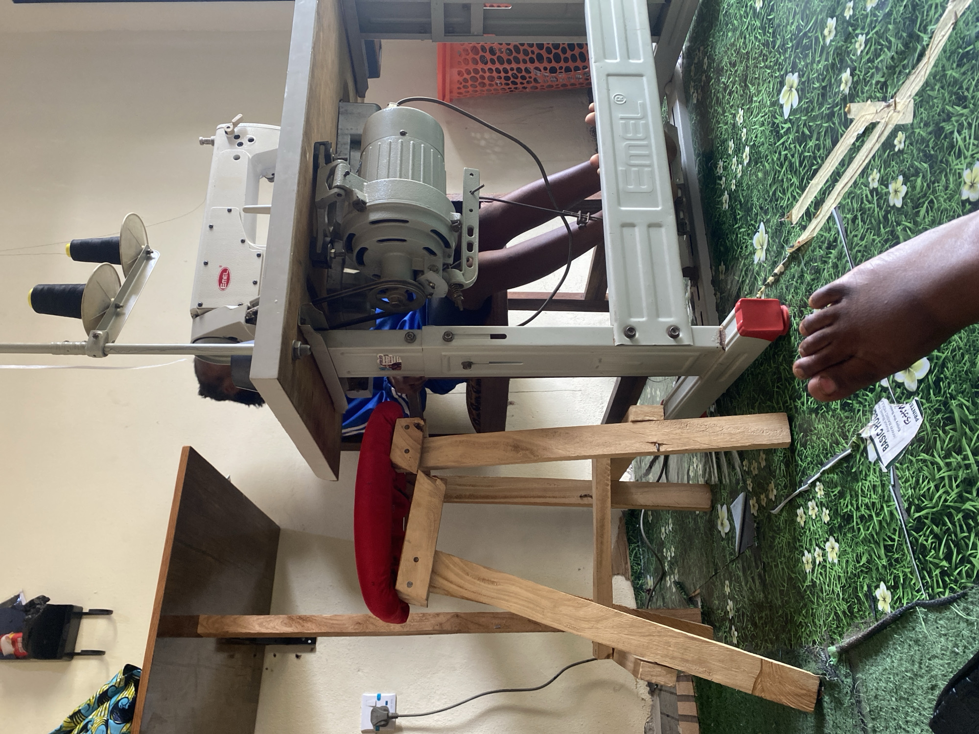 This image depicts a sewing workshop in Jauro Yinu, Nigeria. In the foreground, a person's foot is visible resting on a green floor covered with a pattern of grass and white flowers. Behind it, a wooden stool with a red cushion supports a sewing machine. The sewing machine, a white EMEL brand, is mounted on a metal table. A person, partially obscured, is seated at the sewing machine. Various tools and materials, including spools of black thread, a power cord, and fabric, are present in the workshop. The background reveals a light-colored wall and a wooden shelf with items on it.