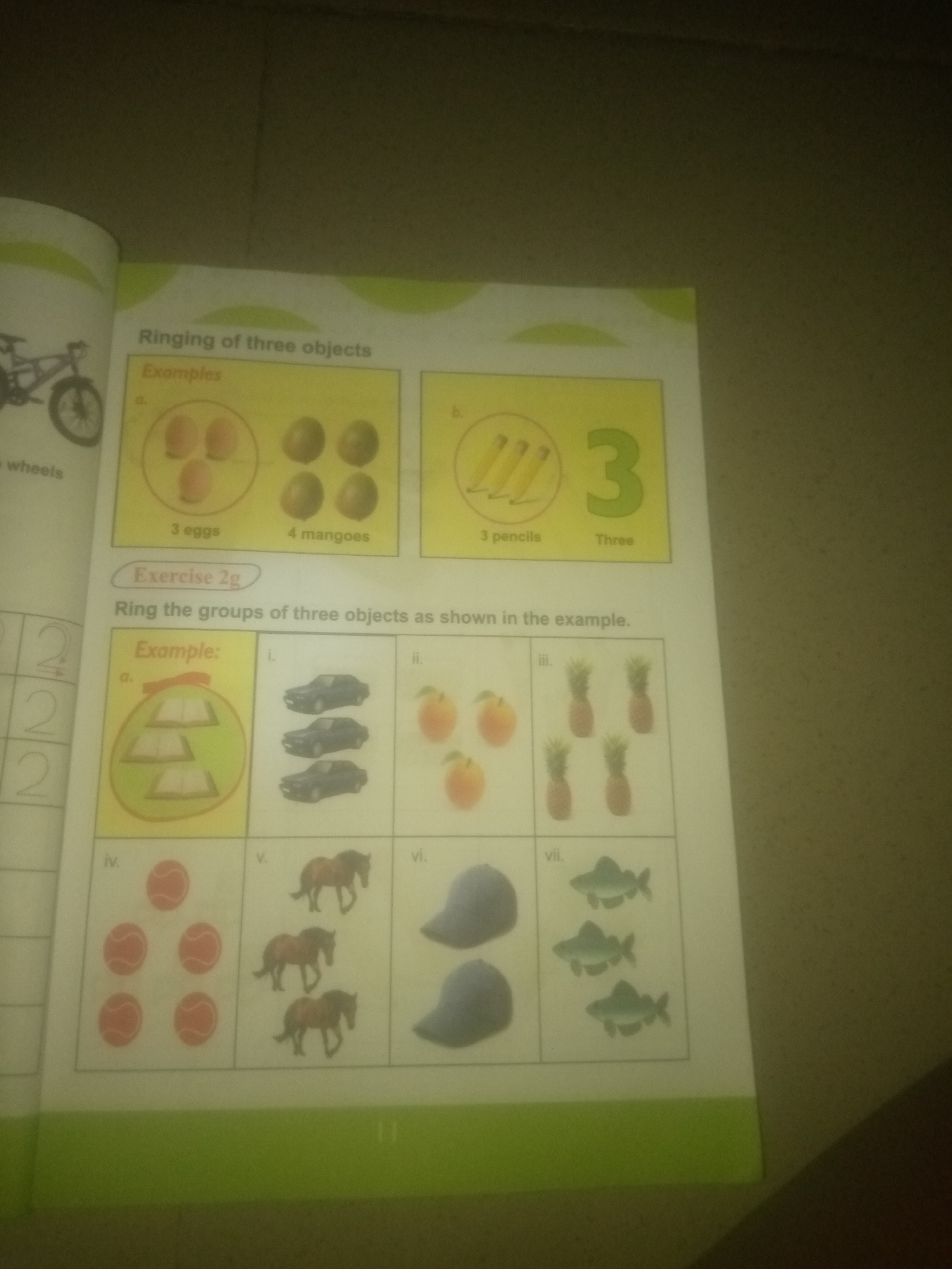 The image shows a page from a children's workbook. The title of the page is "Ringing of three objects". Below this are examples and an exercise.
The examples section has two parts, labeled 'a' and 'b'.
Part 'a' shows a group of three eggs within a circle, and next to it, a group of four mangoes. Below the eggs, it says "3 eggs", and below the mangoes, it says "4 mangoes".
Part 'b' shows three pencils, and next to them, the number "3" and the word "Three". Below the pencils, it says "3 pencils".
The exercise section is titled "Exercise 2g" and instructs "Ring the groups of three objects as shown in the example."
This section contains several items labeled with Roman numerals.
The example for the exercise is labeled 'a' and shows a stack of three books within a green circle.
Following the example, there are items labeled 'i' through 'vii'.
'i' shows three cars.
'ii' shows three peaches.
'iii' shows three pineapples.
'iv' shows five red balls.
'v' shows three horses.
'vi' shows two blue hats.
'vii' shows three fish.
The image is a photograph of a page in a book, taken from a slightly angled perspective, suggesting it is a flat surface such as a table or floor. The background is a muted, textured wall or surface. The image does not contain any information about the city of Jauro Yinu, Nigeria, or any specific geographical location.