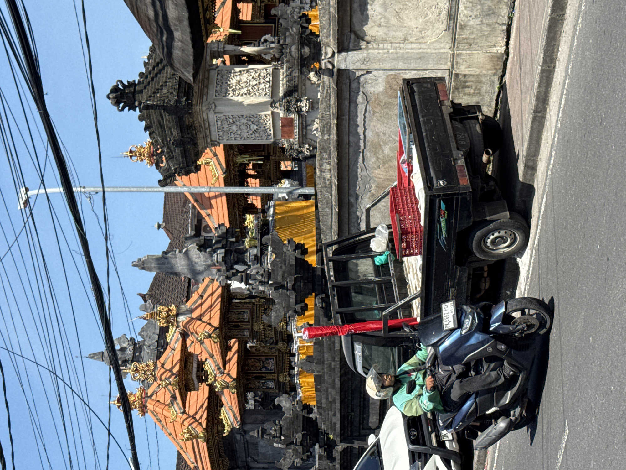 The image captures a street scene in Nusa Dua, Indonesia, featuring a Balinese temple in the background. A black pickup truck with a red cargo net stands next to the temple wall. A blue motorcycle with a rider wearing a helmet and light green jacket is parked beside the truck. A white car is partially visible in the foreground, positioned next to the motorcycle. Overhead power lines are visible against a clear blue sky. The temple displays traditional Balinese architecture with ornate carvings and a tiered roof, decorated with orange and gold details.