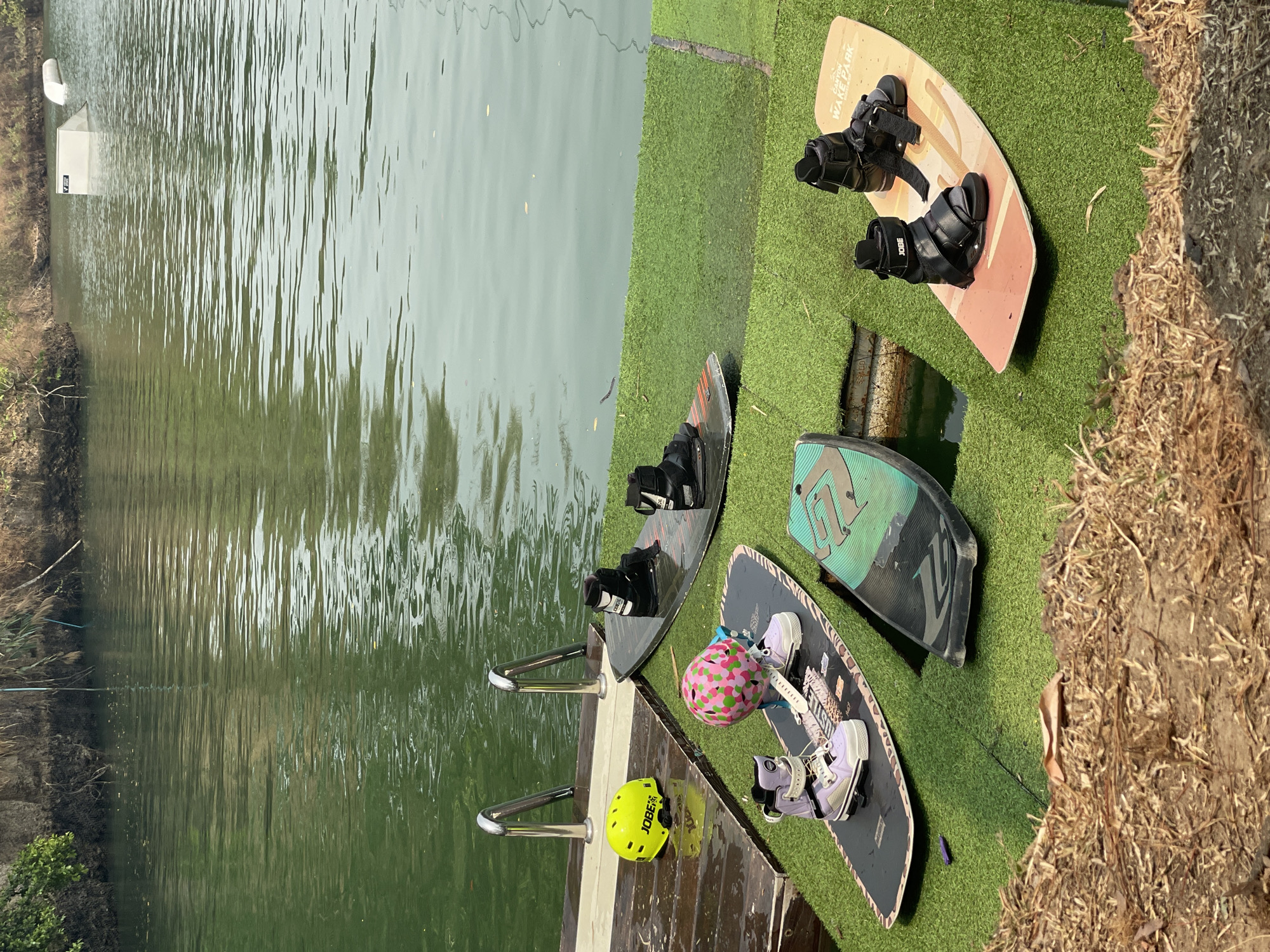 The scene depicts a waterfront area at a wake park or similar recreational facility, likely located in Tambon Nam Phrae, Thailand. The water is calm, with reflections visible on the surface. Several wakeboards are laid out on artificial turf next to the water. These include a light brown wakeboard with black bindings, a black wakeboard with black bindings, a green and black wakeboard, and a dark wakeboard with purple boots and a helmet with a pink floral design. A yellow helmet rests on a wooden platform near the water's edge. Two stainless steel handrails are visible, presumably for entering or exiting the water. The surrounding environment includes vegetation and what appears to be dry grass or straw along the edge of the artificial turf. A structure is visible in the distance across the water. There are no people present in the scene.