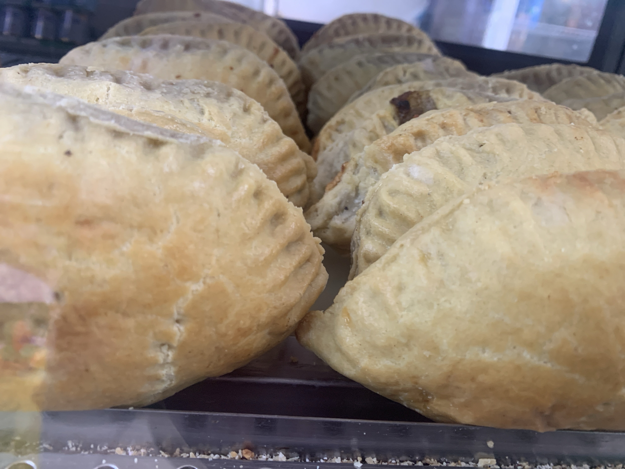 A close-up, eye-level shot displays a tray of golden-brown, crimped turnovers, likely baked goods, arranged in multiple rows. The turnovers are slightly varied in color, with some appearing lighter and others darker. The closest turnovers are in sharp focus, showing detailed texture on their crusts, while those further back are progressively out of focus. The background is blurred, suggesting a commercial setting, possibly a bakery or food display. The turnover in the foreground on the left is the largest and most prominent, with a visible seam along its edge. The turnovers appear to be filled with a dark-colored mixture, visible through small gaps in the crust of some. The lighting is even, highlighting the baked texture of the turnovers. The scene is a still life of food items.