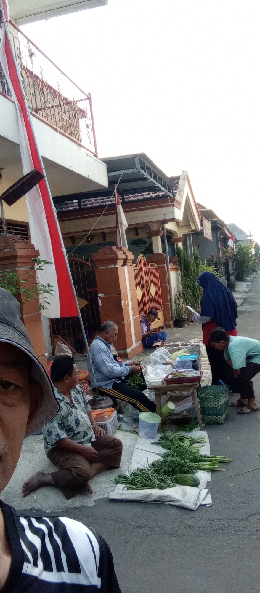 A street scene in Kediri City, Indonesia, shows an outdoor market with several individuals interacting. In the foreground, a person wearing a dark hat and a black and white striped shirt is partially visible, taking the photo from a low angle. To the left, a man sits on the ground, wearing a patterned short-sleeved shirt and brown pants, with his legs crossed. In the middle ground, an older man in a light blue checkered shirt and black pants with yellow stripes sits on a low stool, holding a bunch of leafy greens and appearing to be sorting them. Beside him, a stall displays various vegetables, including long beans, leafy greens, and a pale green gourd. To the right, a person wearing a dark blue hijab and a red undershirt stands, appearing to interact with the vendor. Another individual, a child or young person, in a light blue t-shirt and dark pants, leans over the stall, looking at the produce. In the background, more houses and structures line the street, with Indonesian flags visible, suggesting a public holiday or patriotic display. One person is seated on the ground further back, looking at their phone. The ground is paved with asphalt and concrete, with some debris and plastic bags scattered around. The overall impression is a casual street-side market environment.