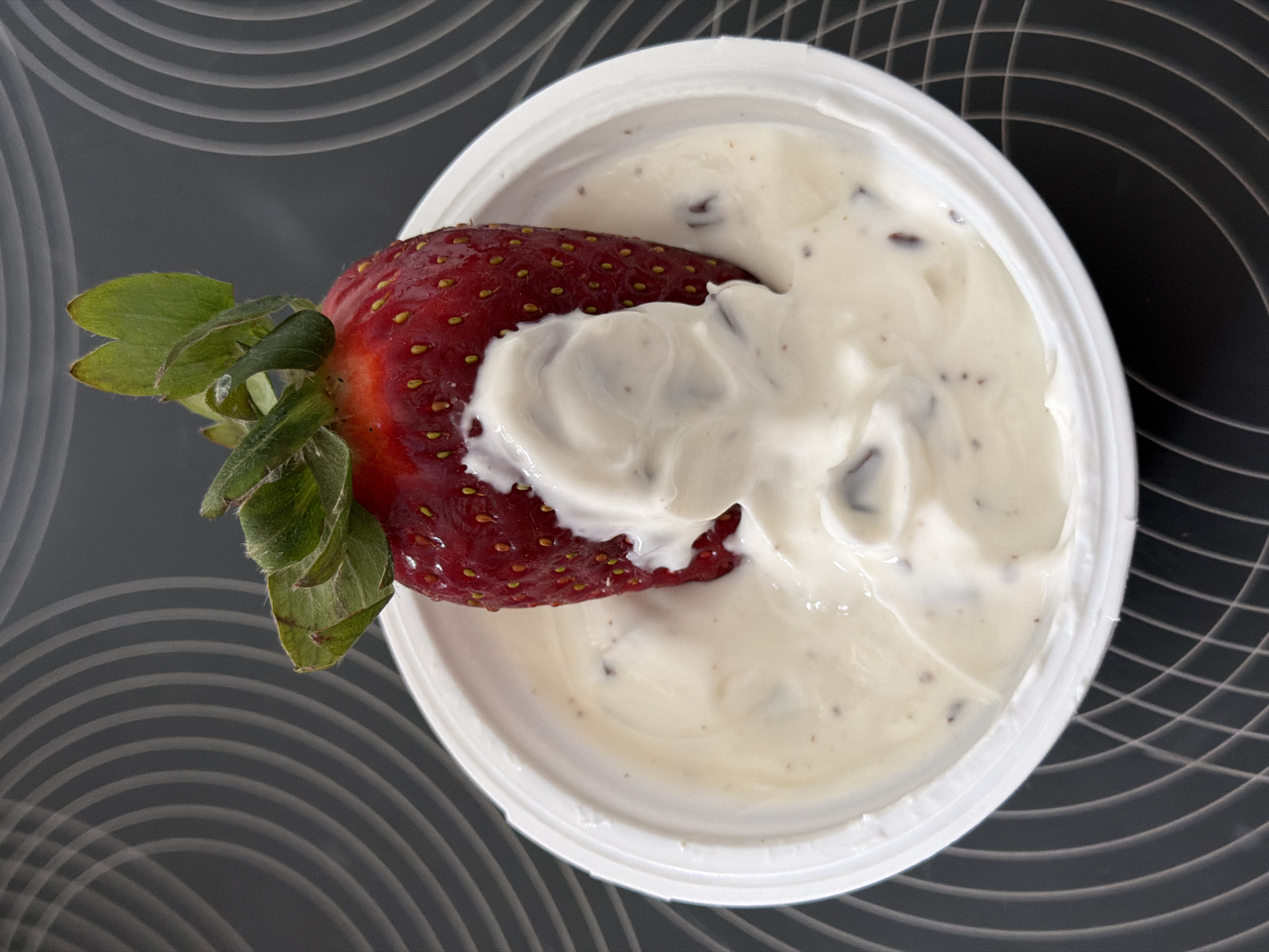 A strawberry with green leaves is dipped in a white, yogurt-like substance inside a white plastic container. The container sits on a surface with a gray, swirling pattern. The scene depicts a snack or dessert. There is no indication of a location in Esch-sur-Alzette, Luxembourg, or any other specific place. There are no people visible in the image.