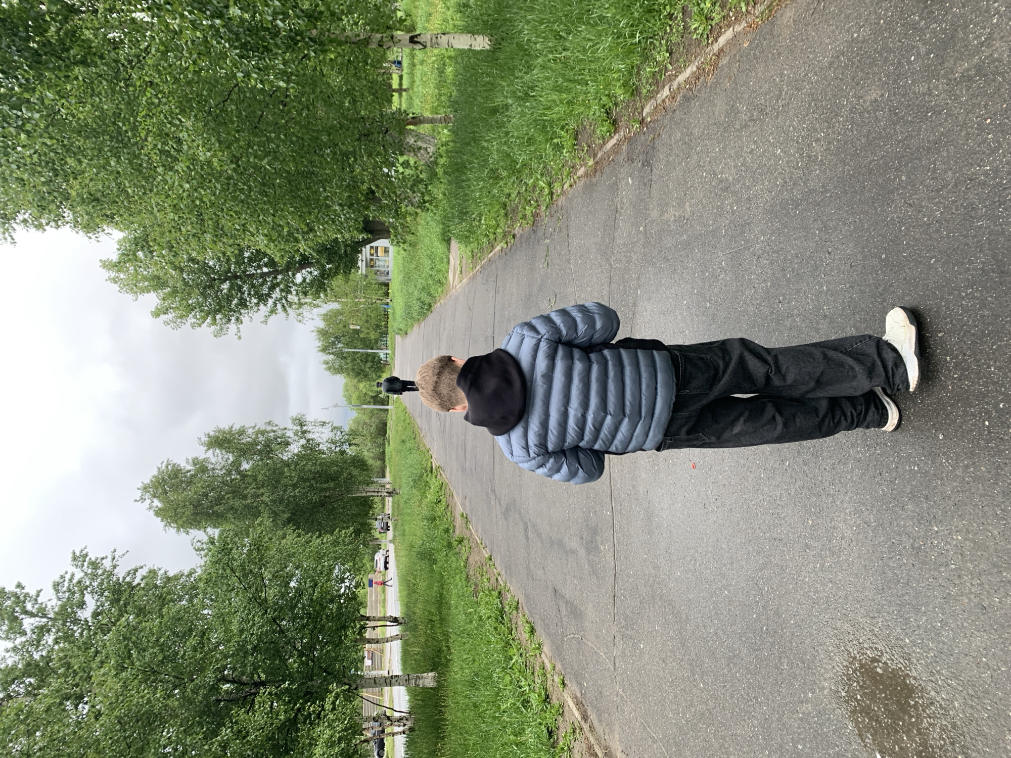 The image, oriented vertically, captures a person from behind, walking or standing on an asphalt path. The individual is wearing a light blue/grey quilted puffer jacket, a black hoodie, black trousers, and white sneakers. The path is damp in areas, suggesting recent rain.
On either side of the path, lush green grass stretches, bordered by numerous trees, primarily birch trees with their distinctive white bark and vibrant green foliage. The sky above is overcast, indicating a cloudy day. In the mid-ground and background, more trees line the path. Distant details include blurred figures of other people, several parked cars, and a small, light-colored kiosk or building with windows on the left side of the path. Further into the distance, there are hints of structures that might be part of a playground or another park amenity.
The activity appears to be a casual outdoor stroll in a park or pedestrian area. The time of day is daylight, under a uniformly grey sky. The scene conveys a calm, everyday atmosphere in what appears to be an urban green space within Severodvinsk, Russia. No visible text is clearly legible in the image.