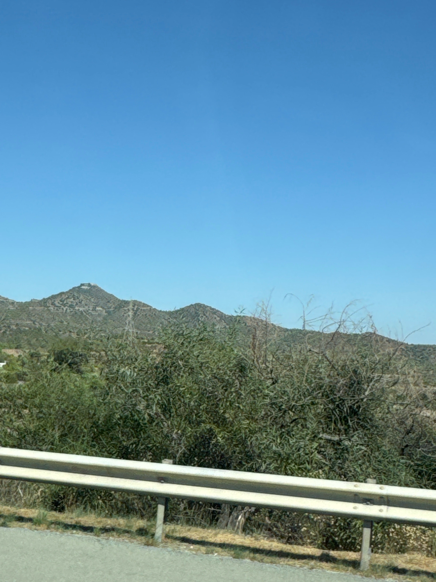The image shows a scenic view of the mountains surrounding Kornos, Cyprus. The sky is a clear, bright blue, and the mountains are covered in sparse, green vegetation. In the foreground, there is a metal guardrail along the side of a road. A tall, metallic structure, possibly a utility pole or tower, is visible on one of the distant hills. The overall impression is one of a natural landscape under a clear sky.
