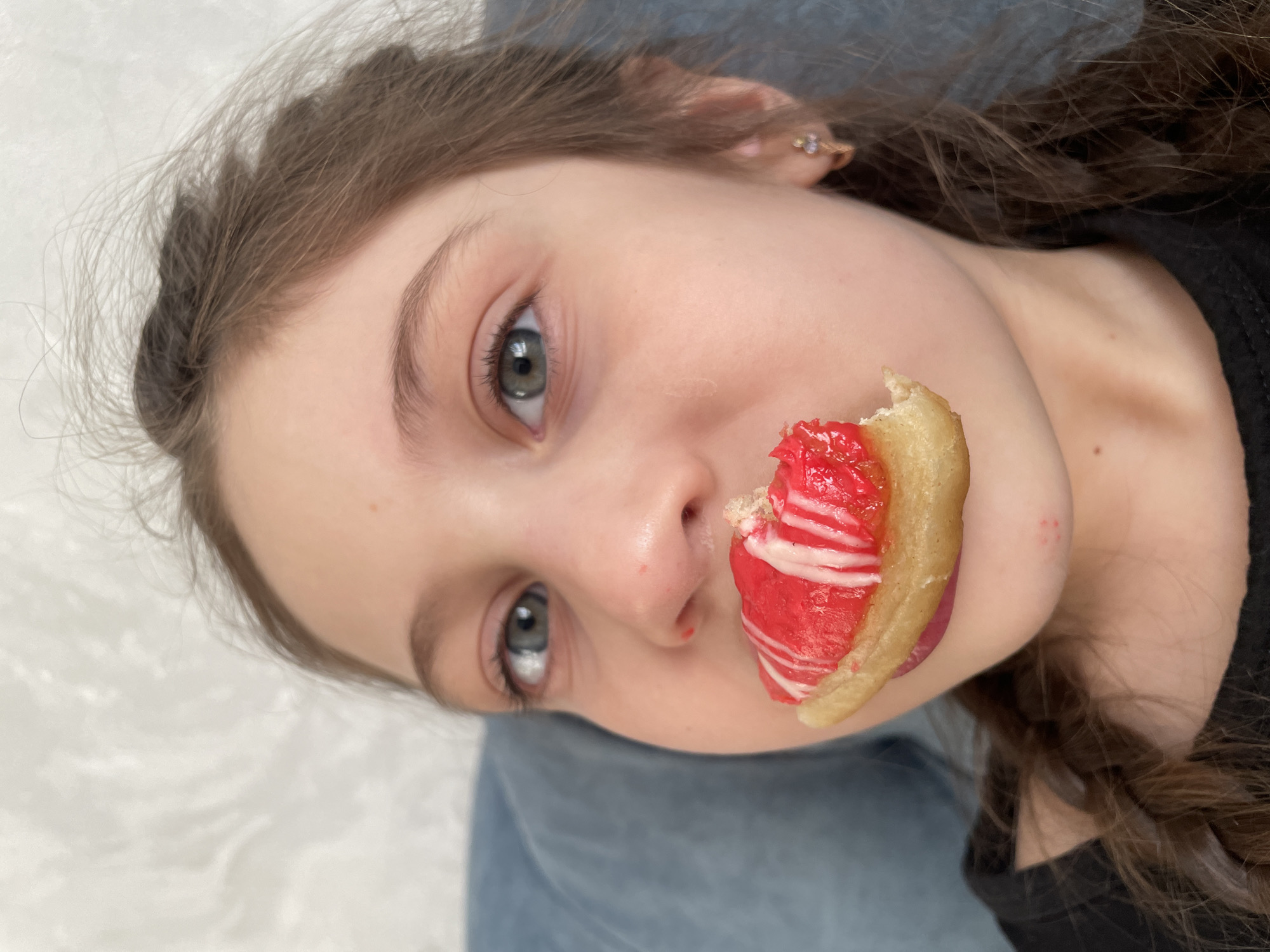 A young girl is lying on her back. She has fair skin, light eyes, and light brown hair that is braided in the back. She has a piece of food, possibly a pastry with red and white filling, near her mouth, and it appears she has taken a bite. She is wearing a dark-colored shirt. The background is blurred but appears to consist of white and blue surfaces, possibly a blanket and clothing. The city is not discernible from this image.
