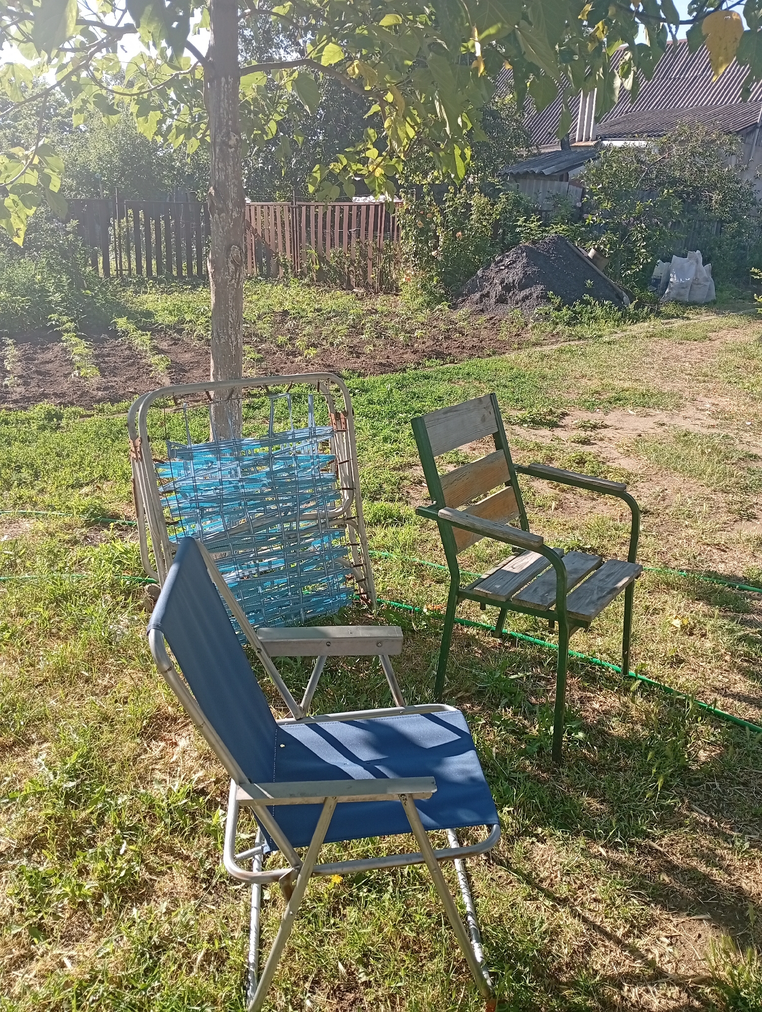 This image depicts a backyard scene in Mykolaiv, Ukraine. In the foreground, a blue canvas folding chair with a silver metal frame sits on a grassy area. To its left and slightly behind, a metal bed frame with a blue plastic mesh is leaning against a tree. To the right of the bed frame, a weathered wooden chair with green metal legs is positioned. The background shows a garden plot, a wooden fence, a pile of dark material, and buildings with corrugated roofs. The overall environment is an outdoor residential area bathed in sunlight. There are no people visible in the image.
