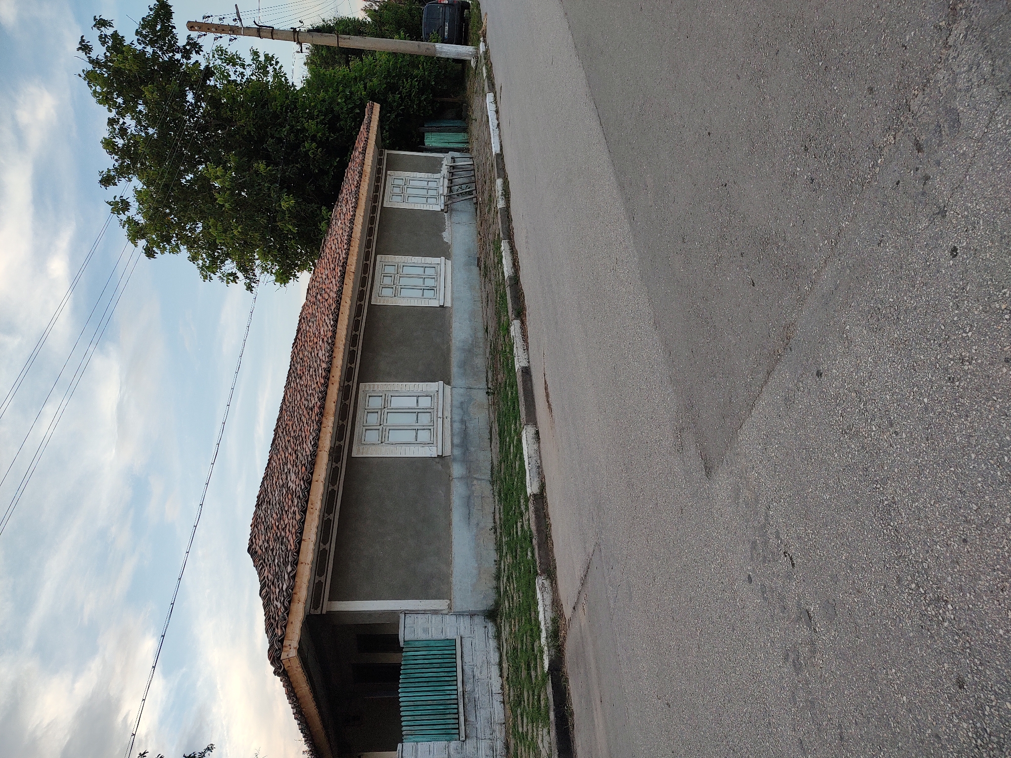 This is a full-frame, eye-level, outdoor shot of a single-story, gray house with a tiled roof. The house is on the left side of the image, viewed from the side, with the front facing away from the viewer. It has three white-framed windows with multiple panes. A green wooden fence stands in front of a section of the house. The house is bordered by a sidewalk with white and black painted curbs, and beyond that is an asphalt road. A large green tree is behind the house on the right, and further back is a utility pole with wires. A dark-colored van is parked on the right side of the road. The sky is partly cloudy with a soft, overcast light. The location is Babuk, Bulgaria.