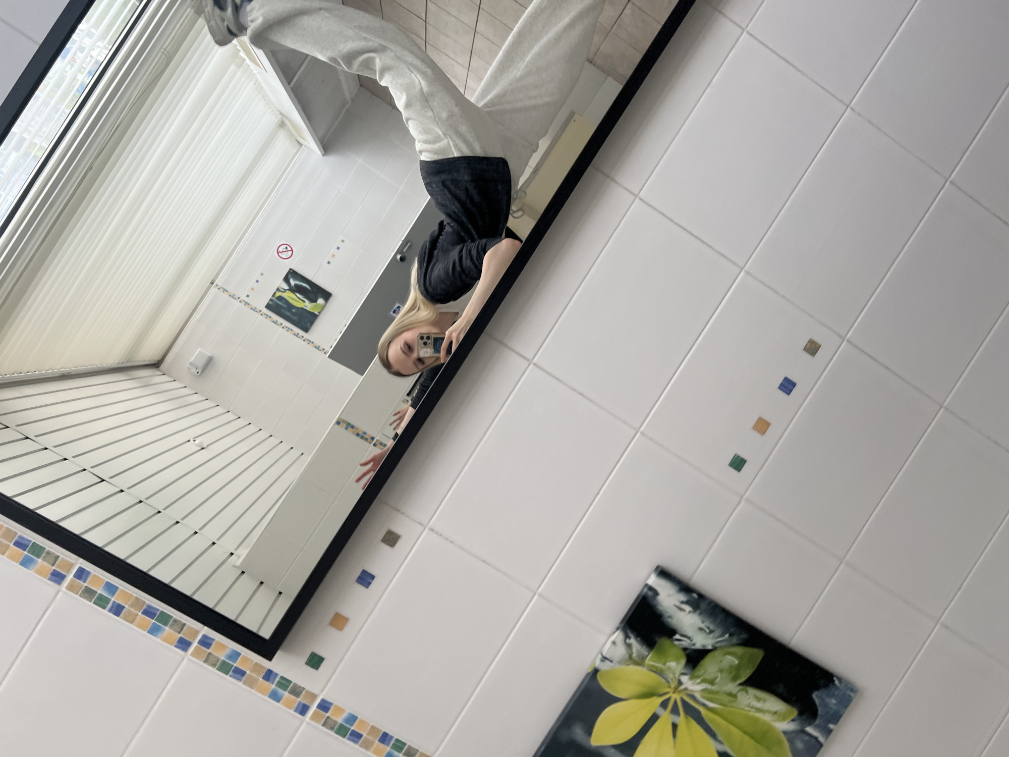 A person is taking a selfie in a bathroom mirror. The reflection shows a window with blinds. The bathroom has white tiles with decorative borders and a picture of a green and yellow leaf. The location is not identifiable as Khimki, Russia.