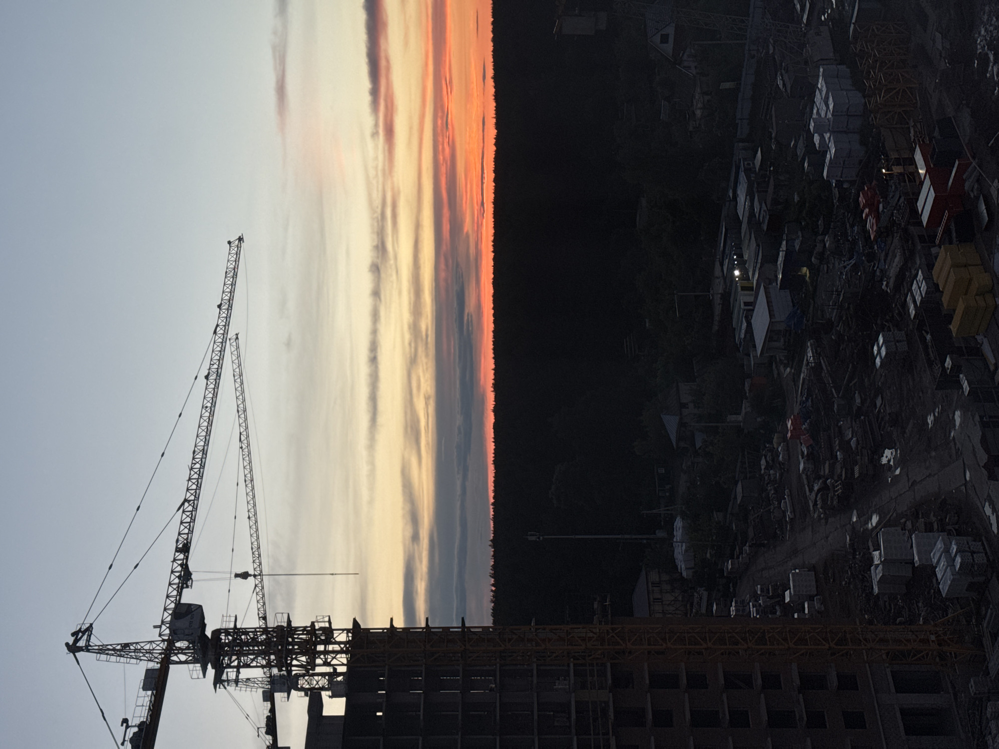 The image depicts a construction site at sunset in Izhevsk, Russia. A tall construction crane dominates the left side, silhouetted against the sky with orange and gray clouds. In the middle, the sky transitions to a dark, dense forest. On the right, a residential area is dimly lit, with visible houses and buildings. Construction materials and equipment are scattered across the site. The sky suggests dusk or dawn.