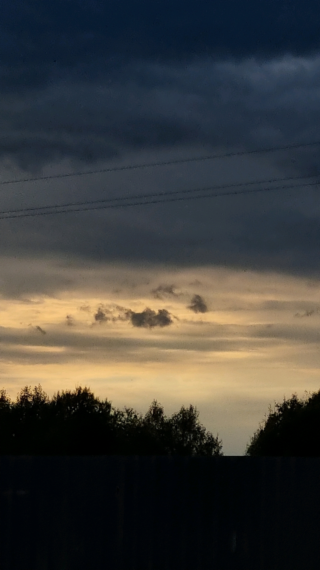 A vertical, eye-level shot captures a dusky sky over the city of Vysokoe, Russia. The sky is divided into two distinct layers. The upper portion is a deep, dark blue, transitioning to lighter shades of gray and yellow near the horizon. Three thin, dark horizontal lines, likely power lines, are visible across the upper part of the sky.
Below the power lines, the sky brightens to a warm yellow-orange hue, indicating either sunrise or sunset. A few small, dark gray clouds are scattered within this lighter sky. The foreground is dominated by a silhouetted line of trees, forming a dark, uneven edge against the lighter sky. Beneath the trees, a dark, rectangular shape suggests a fence or structure, further obscuring the ground. The image appears to be taken during twilight hours, with the setting sun casting a soft glow. There are no visible people or discernible actions taking place in the scene.