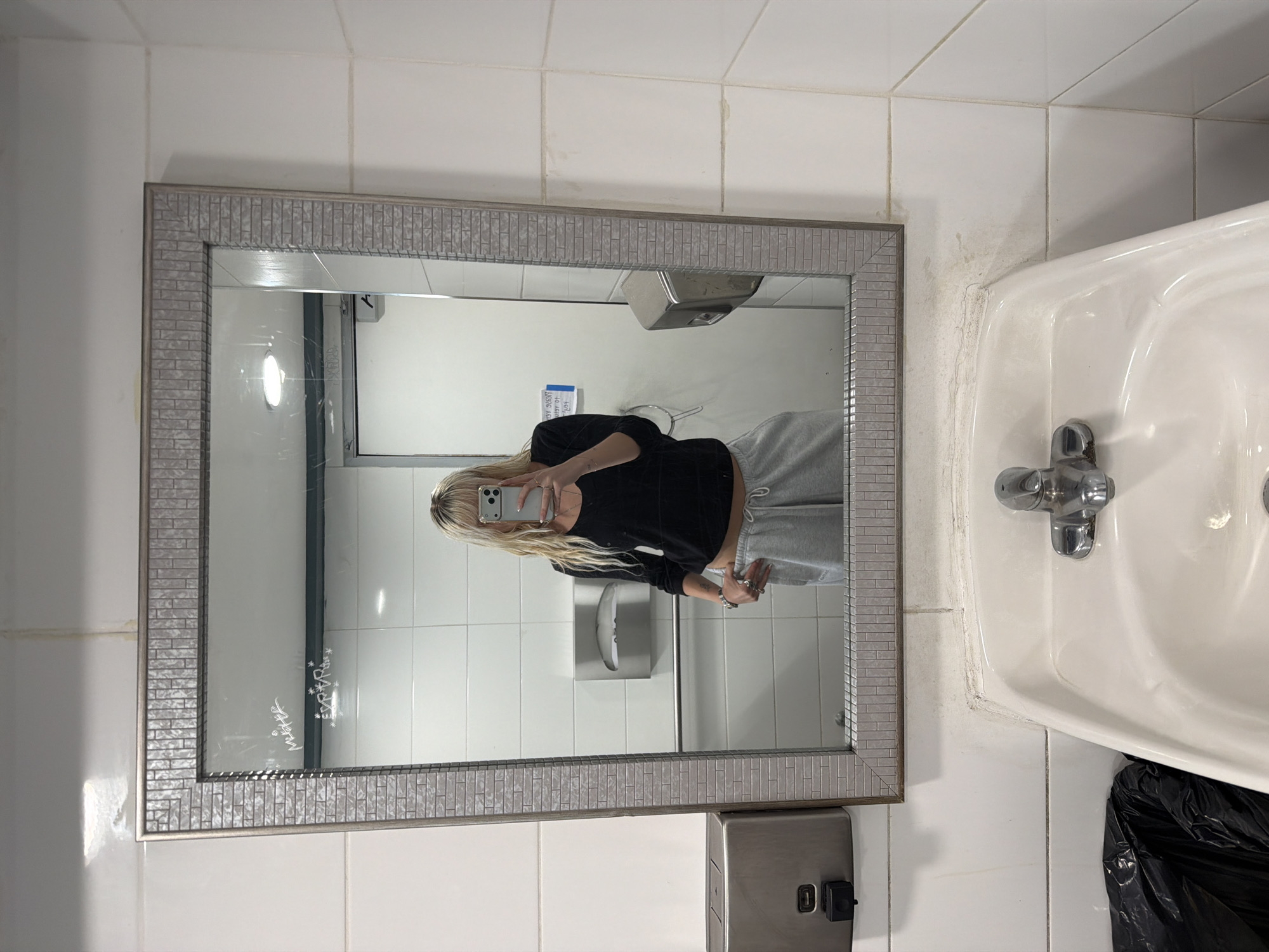 An upward-facing, eye-level shot captures a person taking a mirror selfie in what appears to be a public restroom. The central subject is a person with long blonde hair, wearing a black long-sleeved shirt and light grey sweatpants, holding a smartphone to capture their reflection. Their face is partially obscured by the phone.
The mirror, framed with a decorative silver-toned mosaic pattern, is mounted on a white tiled wall. Inside the mirror's reflection, behind the person, a paper towel dispenser and a hand dryer are visible. A blue sign, partially legible, is also reflected in the background.
To the right of the mirror, a white ceramic sink with a chrome faucet is prominent. A black trash bag rests beneath the sink. The white tiled walls show signs of wear and discoloration in the grout lines, particularly around the edges of the mirror and the sink, suggesting an older or well-used facility.
Notable details include text scratched onto the bottom left corner of the mirror, which reads "MALCHE" and "EVA" followed by stars and other symbols. The overall impression is a candid moment captured in a functional, possibly aged, restroom environment.
