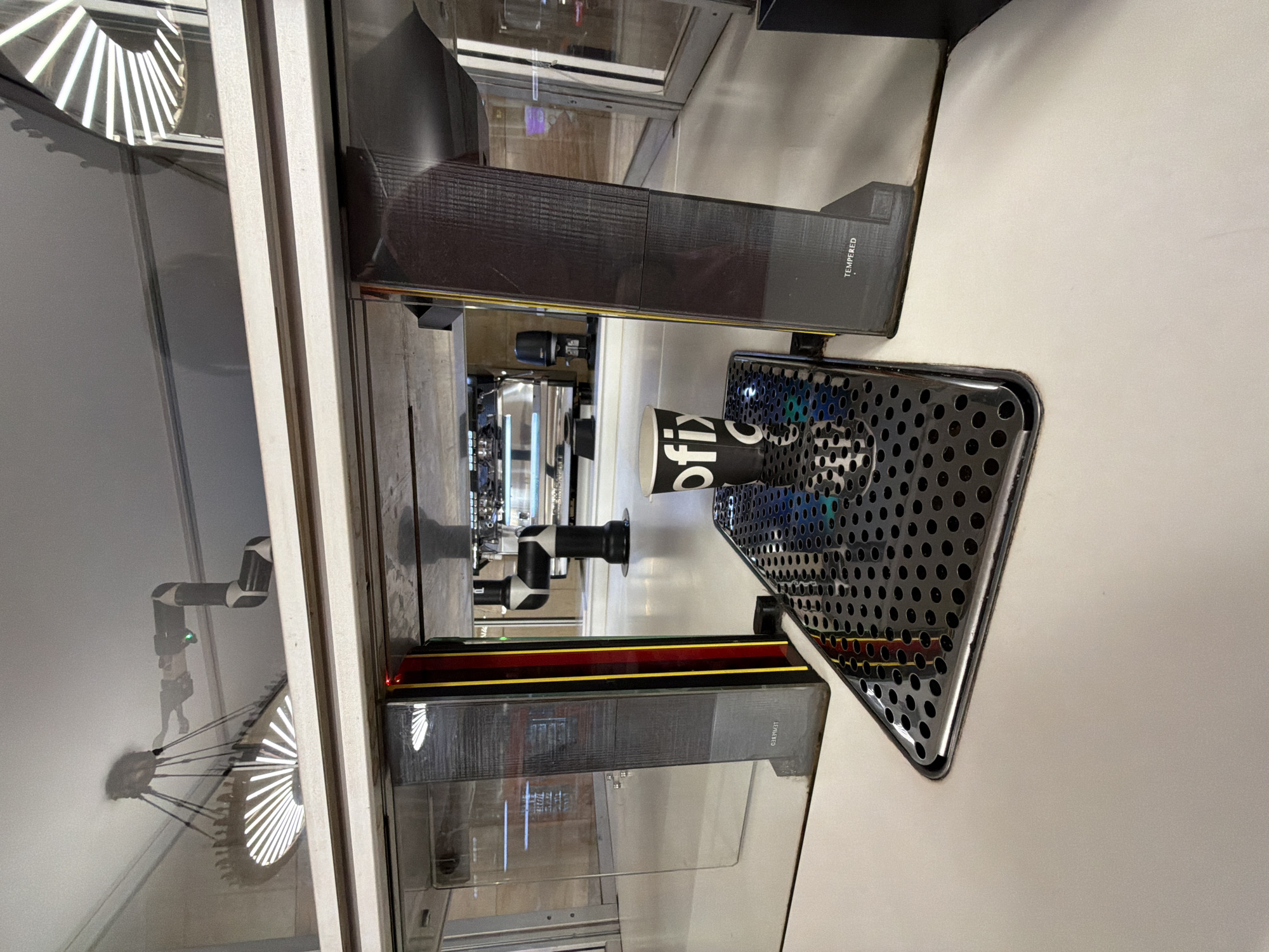 A sideways interior shot depicts a coffee shop, likely in Moscow, Russia, given the Cyrillic branding on a coffee cup. The image features a tempered glass partition, behind which is a coffee machine. A metal grate is visible, and on the white counter, a black and white "ofix" branded paper cup is placed. The reflection of a lamp is visible in the glass partition.