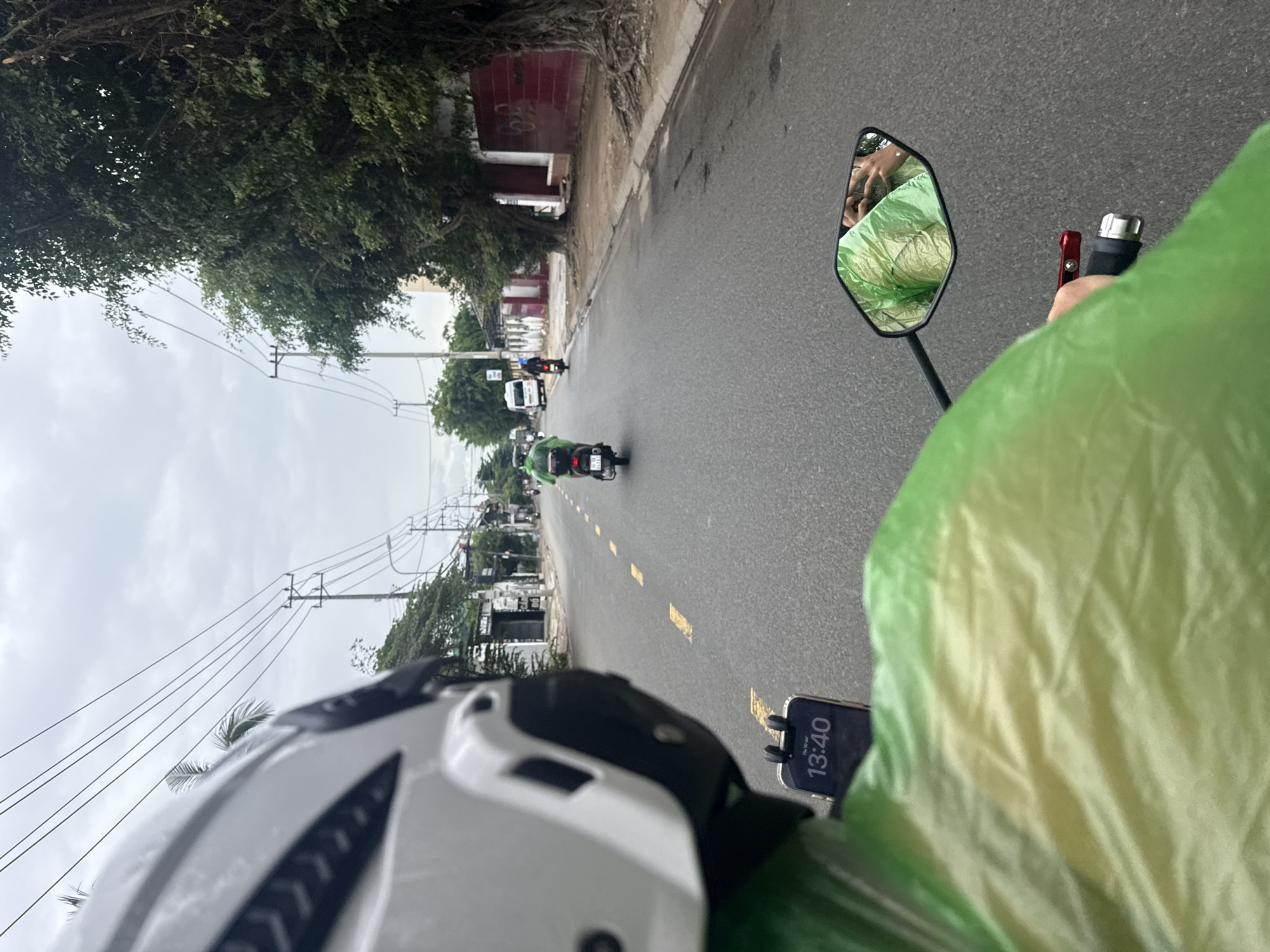 In a street scene in Nha Trang, Vietnam, the point of view is from a person riding a scooter, indicated by the handlebars and side mirror. The rider is wearing a white helmet and a bright green, possibly reflective, rain jacket. A smartphone is mounted on the handlebars, displaying the time as 13:40. The road is asphalt with yellow dashed lines, suggesting it is a two-lane street. In front of the rider, another scooter with a person in a similar green jacket is visible, also heading in the same direction. Further down the road, a white van and another scooter are present. The environment includes buildings and trees along the right side of the street, with power lines overhead. The sky is overcast, indicating potentially gloomy weather. The overall impression is of a common urban commute.