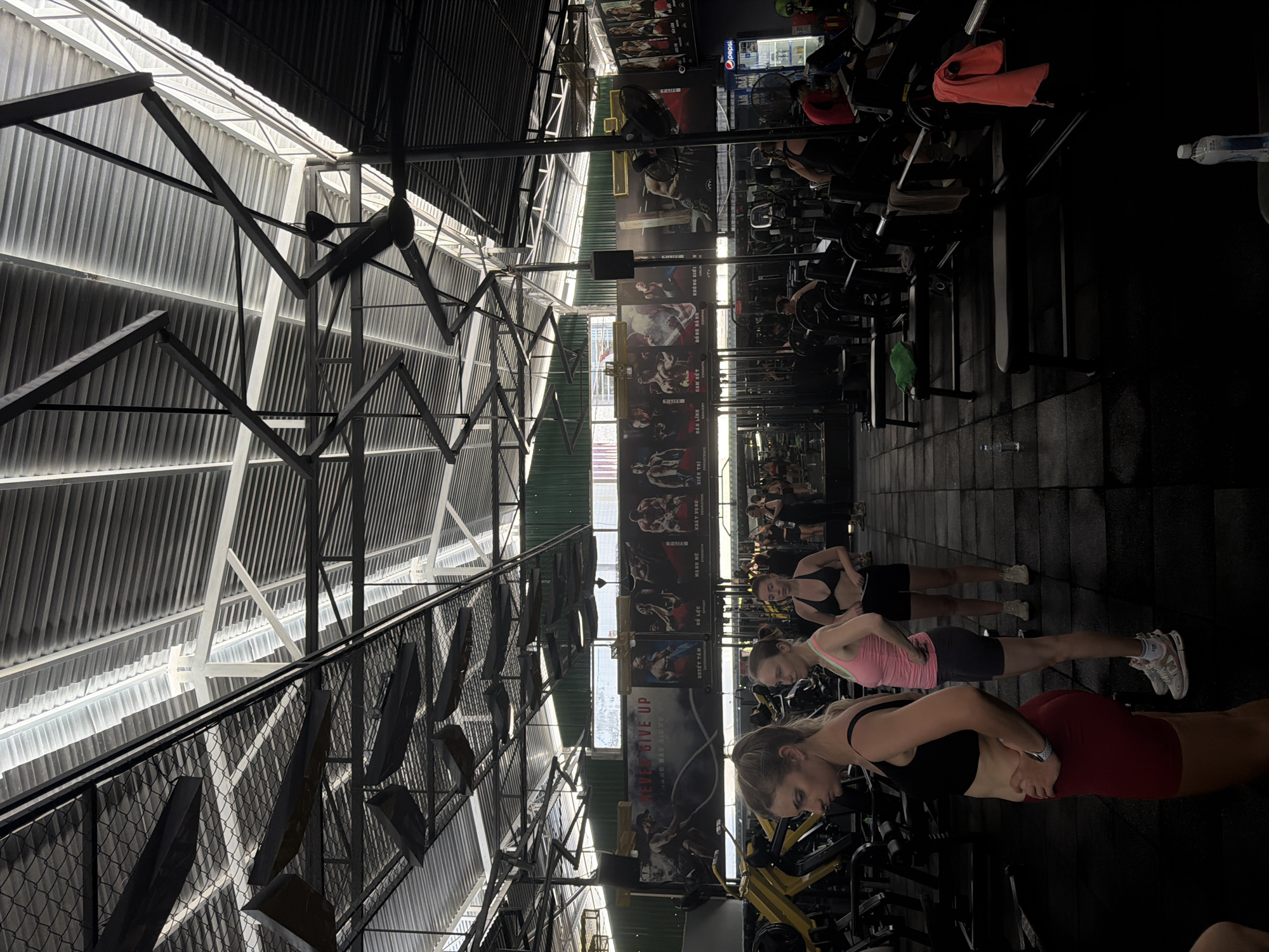 The image depicts the interior of a dimly lit gym, likely located in Bac Nha Trang Ward, Vietnam, as suggested by Vietnamese text on posters. The ceiling is industrial, with exposed metal beams and corrugated roofing, punctuated by fluorescent lighting and large ceiling fans. In the foreground, three women are visible. The woman on the left wears a black sports bra and red shorts, with her hands on her hips. To her right stands a woman in a pink tank top and grey shorts. Further to the right, another woman is dressed in a black sports bra and black shorts.
The gym appears to be equipped with various weightlifting machines and benches, some of which are occupied by other individuals engaged in exercise or resting. Prominent posters with motivational imagery and Vietnamese text adorn the back wall. To the right, a refrigerated display case, possibly containing beverages, is partially visible, with a Pepsi logo on it. The overall atmosphere suggests an active fitness environment.