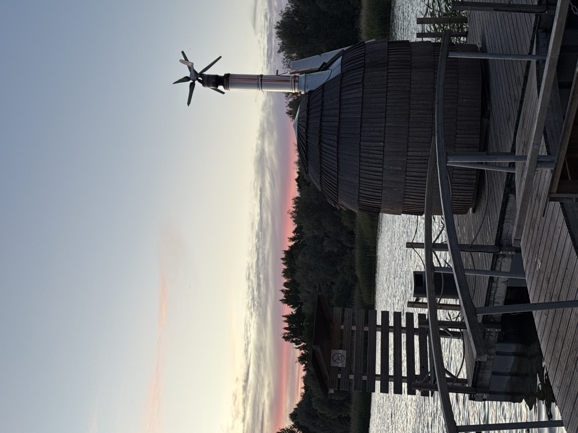 The image depicts a wooden sauna structure on a pier at sunset. The sauna is barrel-shaped and dark brown, with a conical roof and a metallic chimney topped with a weather vane. To the left of the sauna is a wooden structure with slats and a roof, resembling an outhouse or changing room, with a visible "no smoking" sign. The pier is made of weathered wooden planks and features a railing. The scene is set against a body of water with gentle ripples. In the background, a dense line of dark green trees forms the horizon, and the sky displays a soft gradient of pale blue to pink, indicative of twilight. The location is identified as Tallinn, Estonia. There are no people visible in the image.