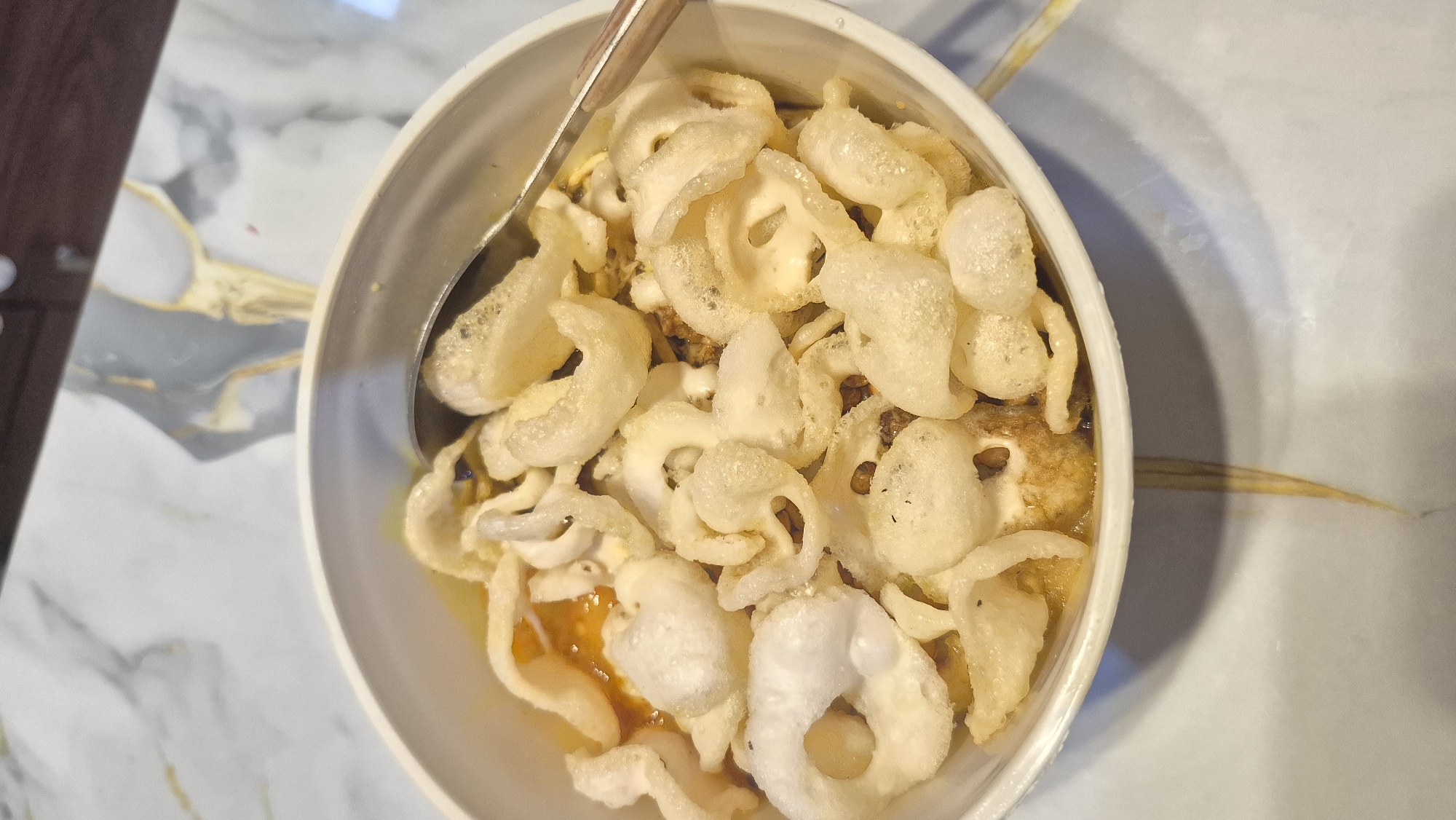 A close-up, top-down view shows a white bowl filled with a dish of yellow broth and topped with numerous light-colored, puffy fried crisps. A metal spoon rests in the bowl, partially submerged in the broth. The bowl is placed on a surface with a marble-like pattern, featuring grey and white swirls with thin yellow lines. In the background, the surface continues, with a dark brown wooden element visible at the very top. The overall impression is of a food presentation, likely in a restaurant or home setting within South Tangerang, Indonesia.