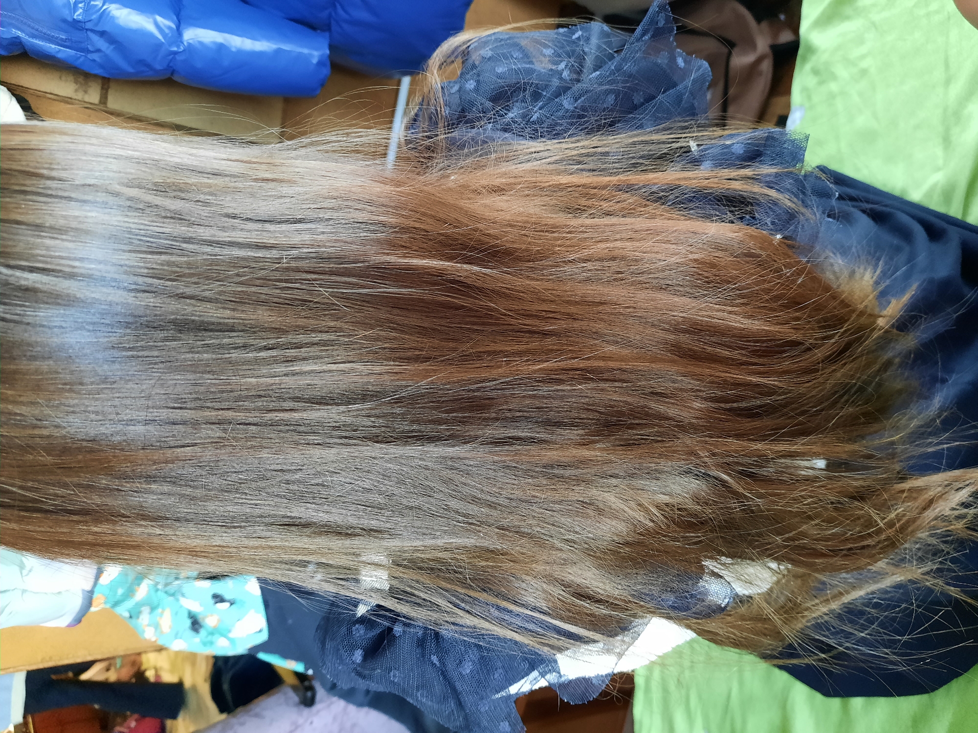 The image shows a section of long hair, transitioning from a darker, possibly dyed brown at the root to a lighter, reddish-brown at the ends. The hair is lying on a dark, sheer fabric with polka dot details, which is partially resting on a light green surface. In the periphery of the frame, there are glimpses of other objects: a blue, shiny material possibly a jacket, and an area with fabric featuring a light blue patterned print. The photograph was taken in Krasnogorskoye, Russia.