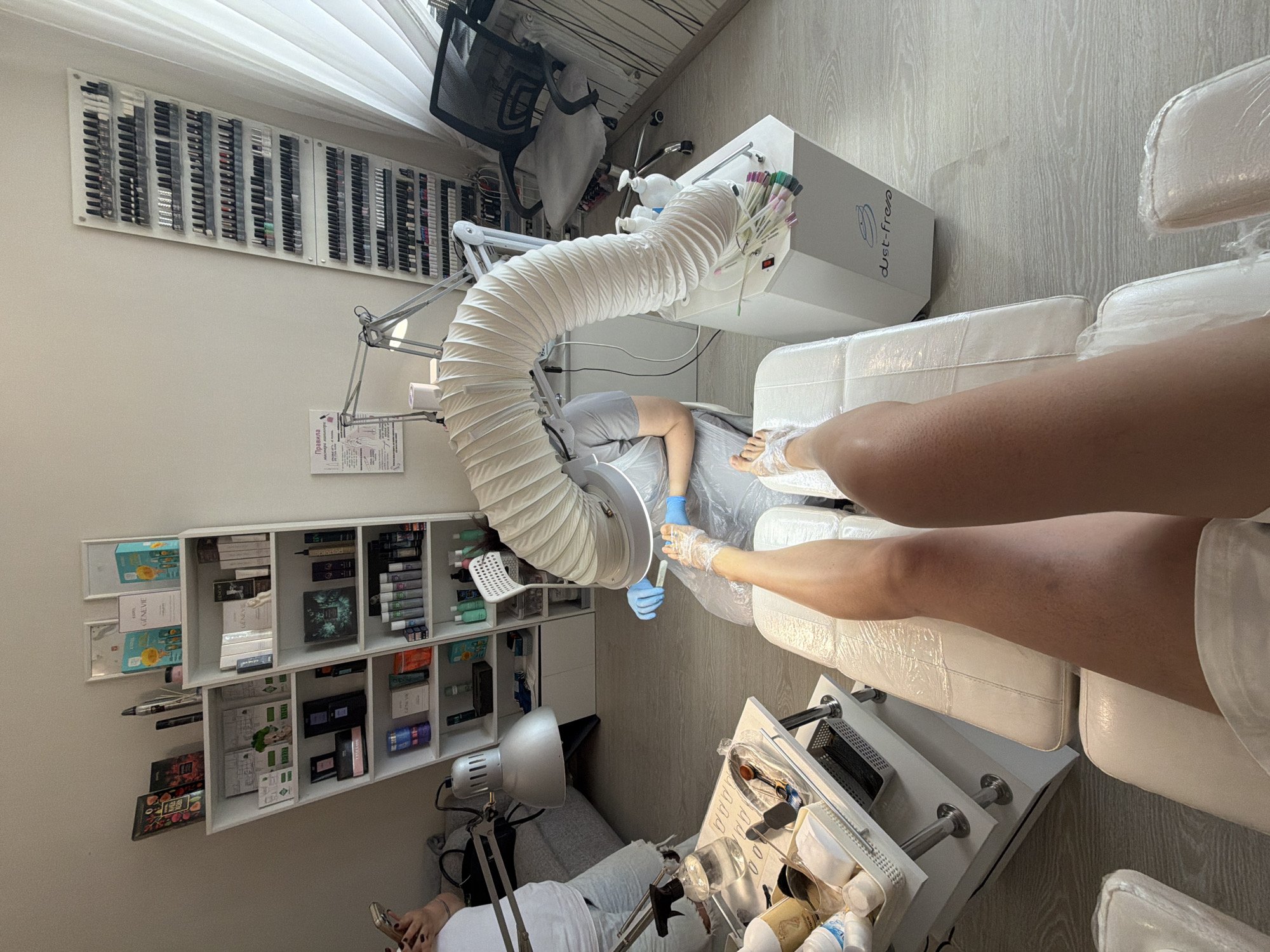 In a salon located in Birobidzhan, Russia, a pedicure is being performed. A client, with legs covered in plastic wrap, is seated on a white pedicure chair. A technician, wearing blue gloves and a grey apron, is meticulously working on the client's foot. The technician is using a dust collector with a flexible hose connected to a white base unit labeled "dust-free". Various nail polish samples are displayed on the base unit. To the left, a trolley is stocked with bottles, jars, and tools, including a spray bottle of water. In the background, shelves are filled with cosmetic products and nail polish bottles. A mesh-backed chair and a large window with sheer curtains are also visible.